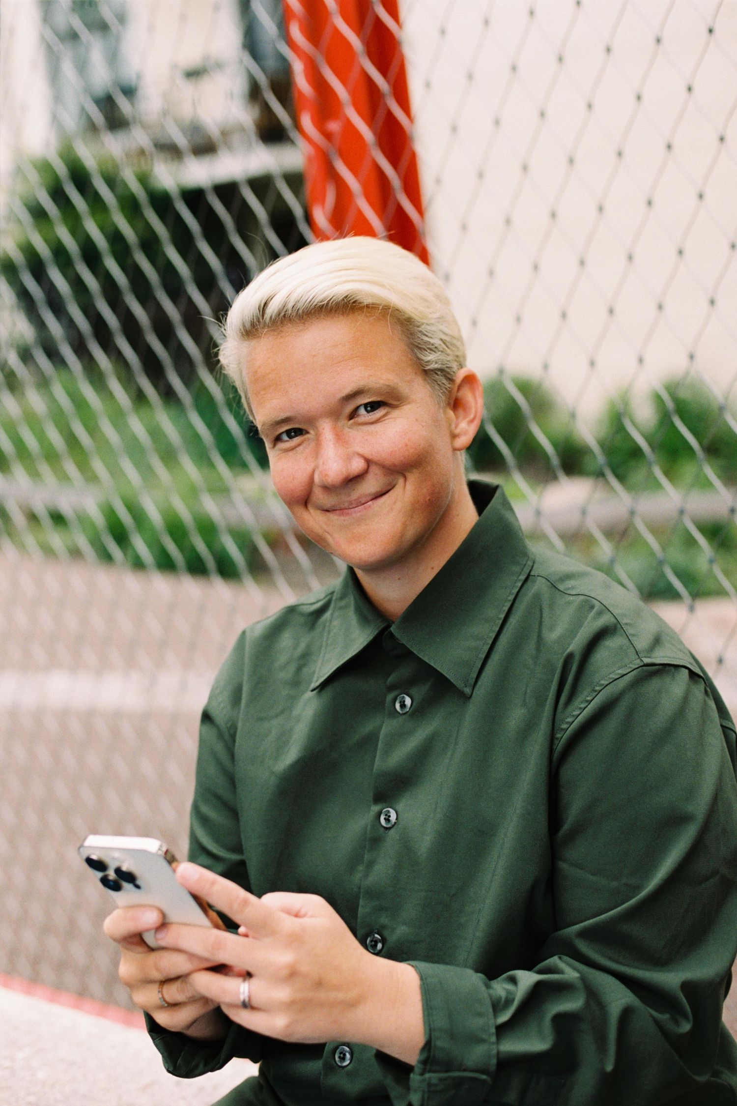 Person in green button-up shirt smiling while using a mobile phone outdoors by a chain link fence.