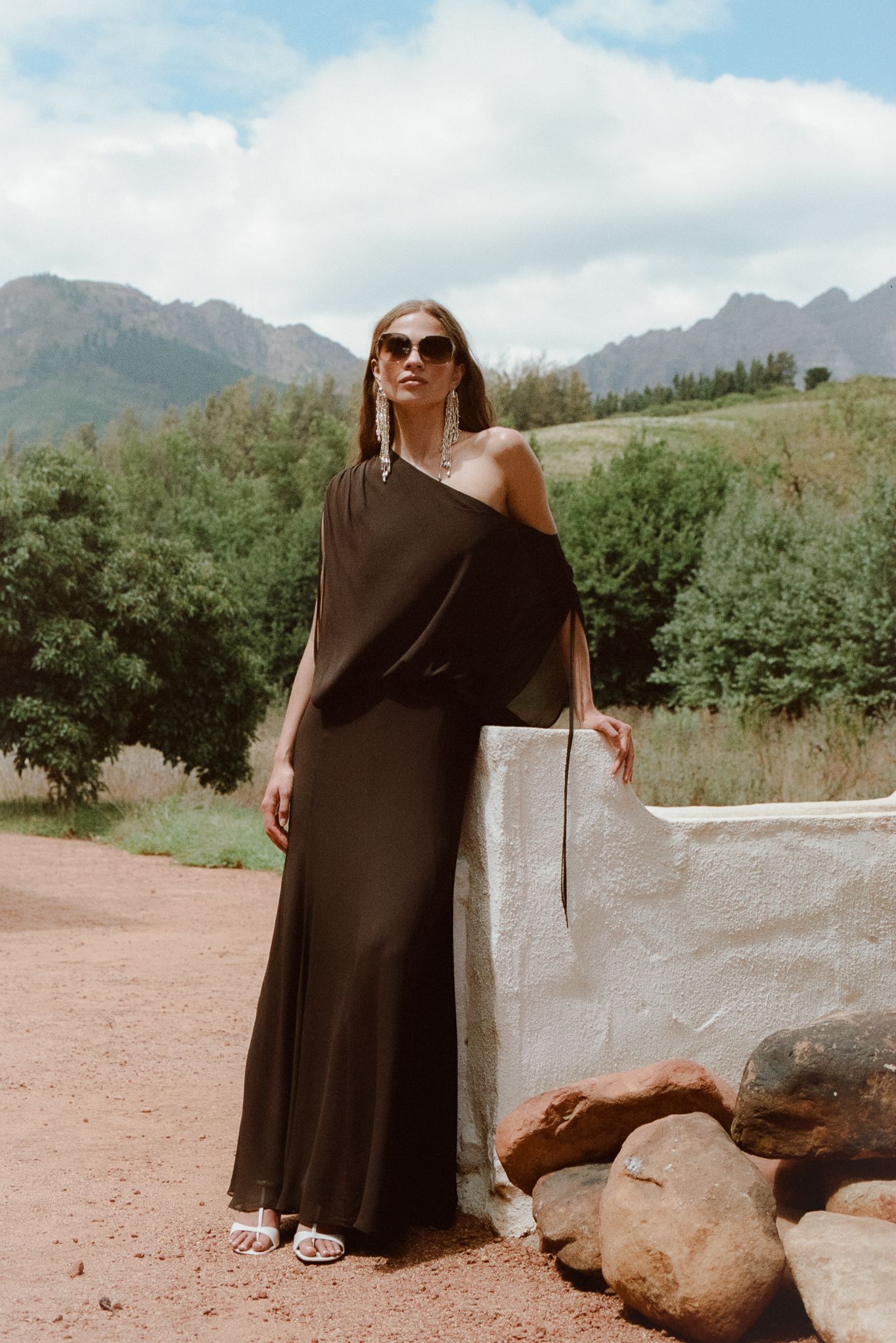 Woman in black one-shoulder maxi dress and sunglasses poses near rocks with mountain landscape backdrop.