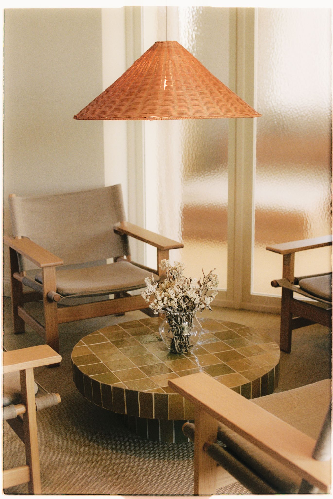 Mid-century modern living room with wicker pendant lamp, tiled coffee table, wooden armchairs, and dried floral arrangement.