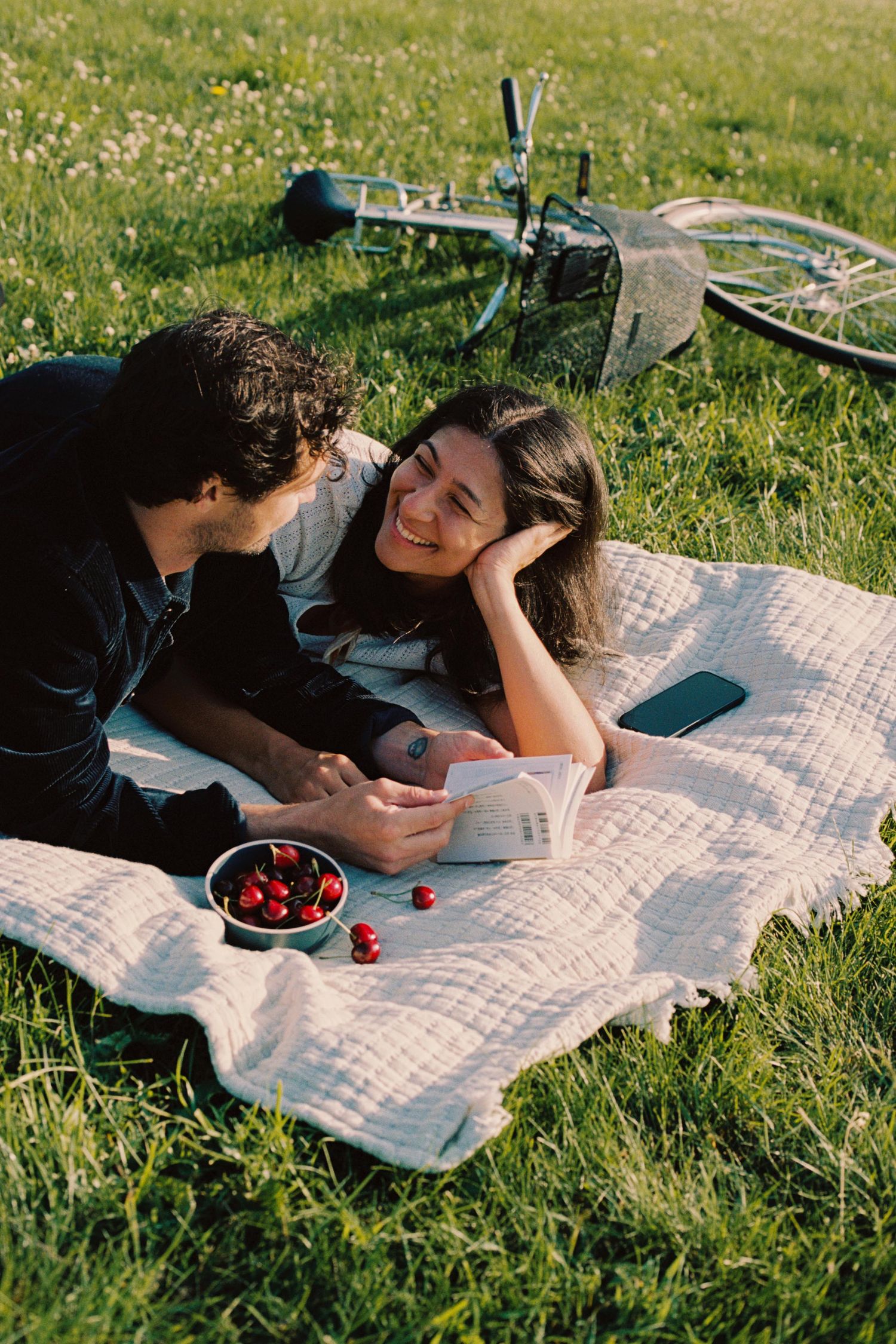 A couple relaxes on a picnic blanket in the grass while reading books on a sunny summer day.