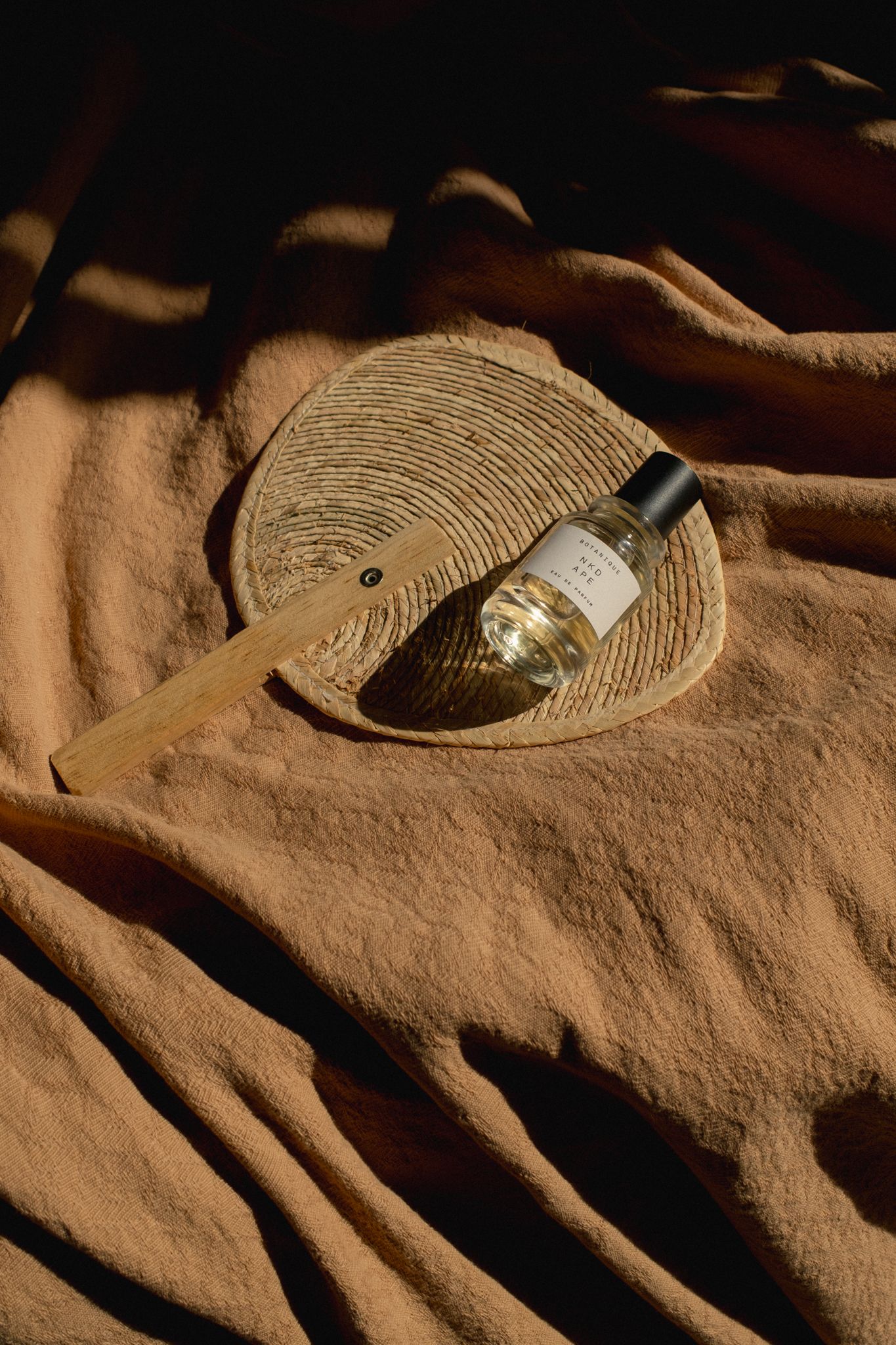 A fan made from natural materials rests on a camel-colored blanket, topped with a minimalist perfume bottle by Naken Ape.
