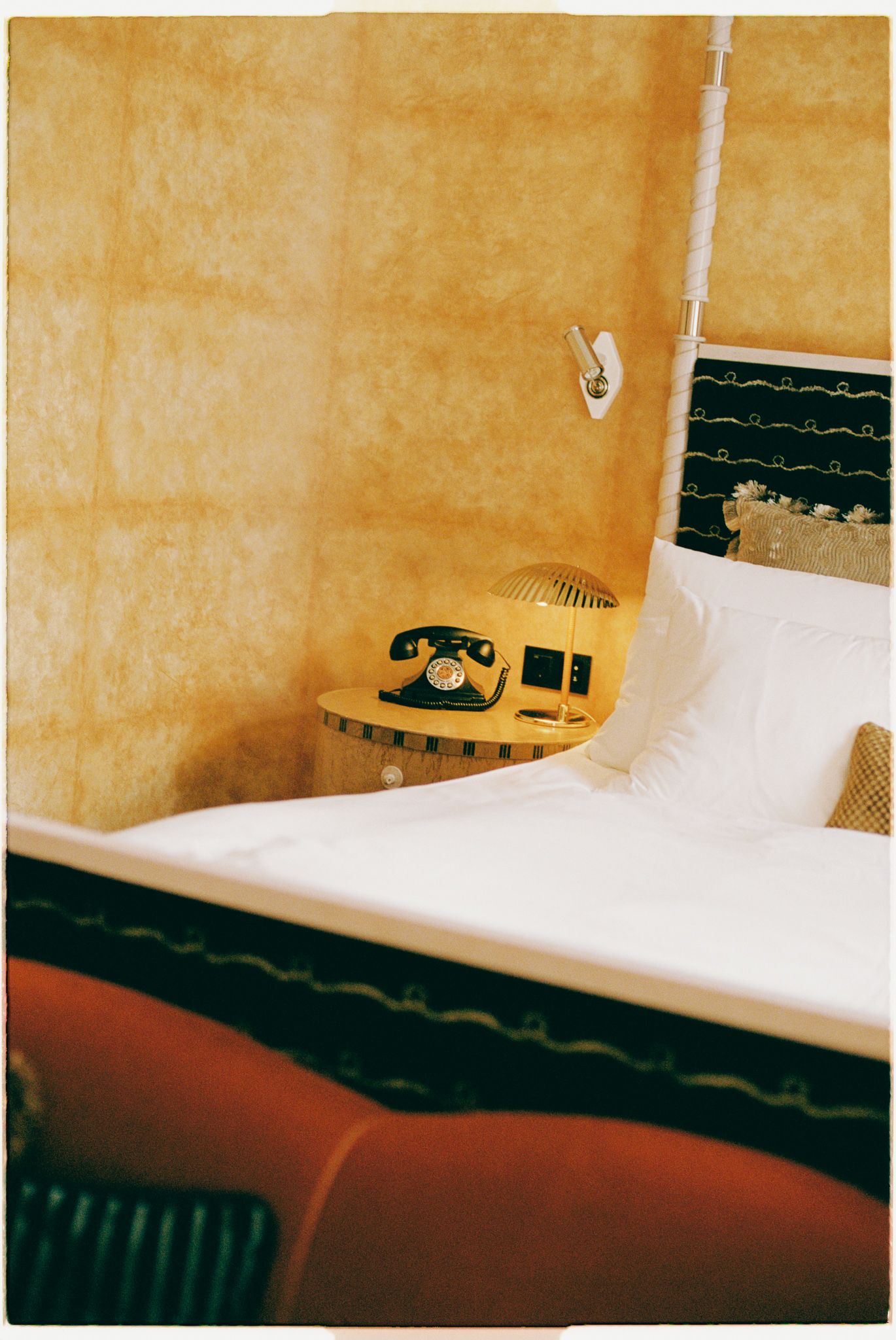 A vintage yellow rotary telephone sits on a white surface next to a dark patterned cushion in a retro-styled room.