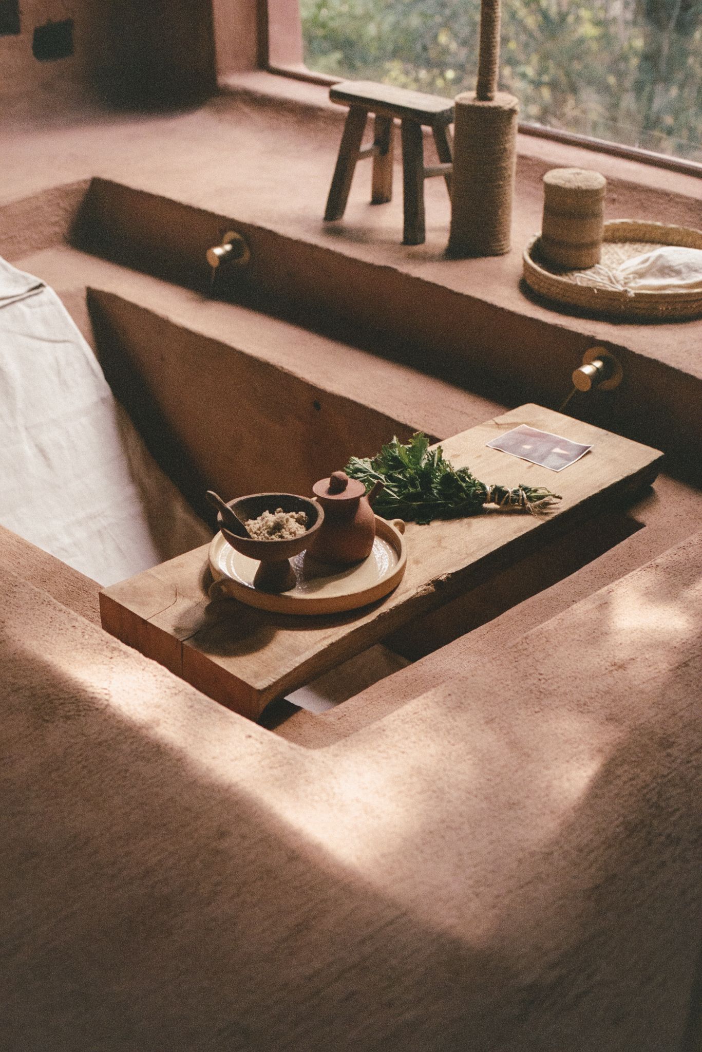 Rustic concrete bathtub with wooden tray holding herbs, clay bowls, and a book in warm natural light.