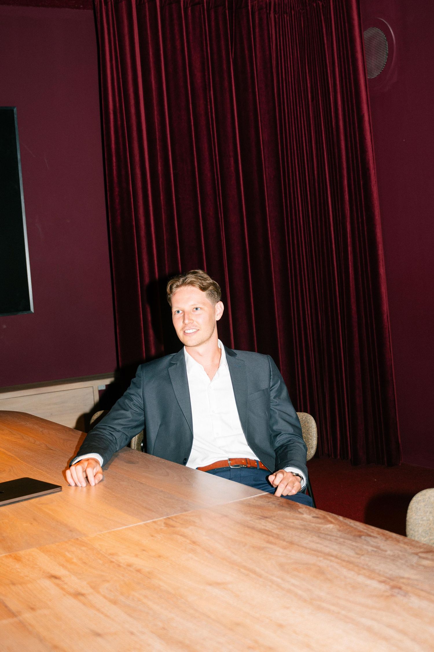 Professional seated at wooden conference table in business attire against red curtain backdrop.