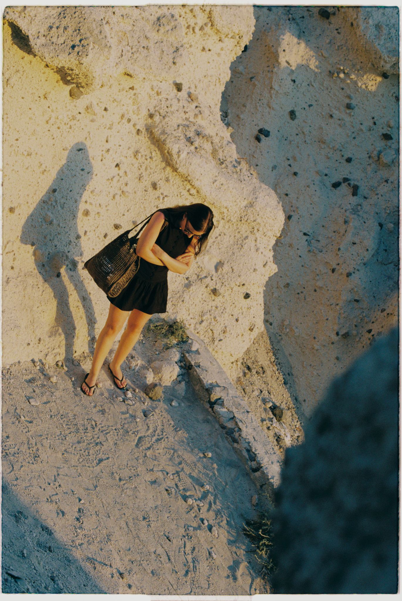 Someone in a black dress stands on a rocky cliff face casting a long shadow during golden hour.