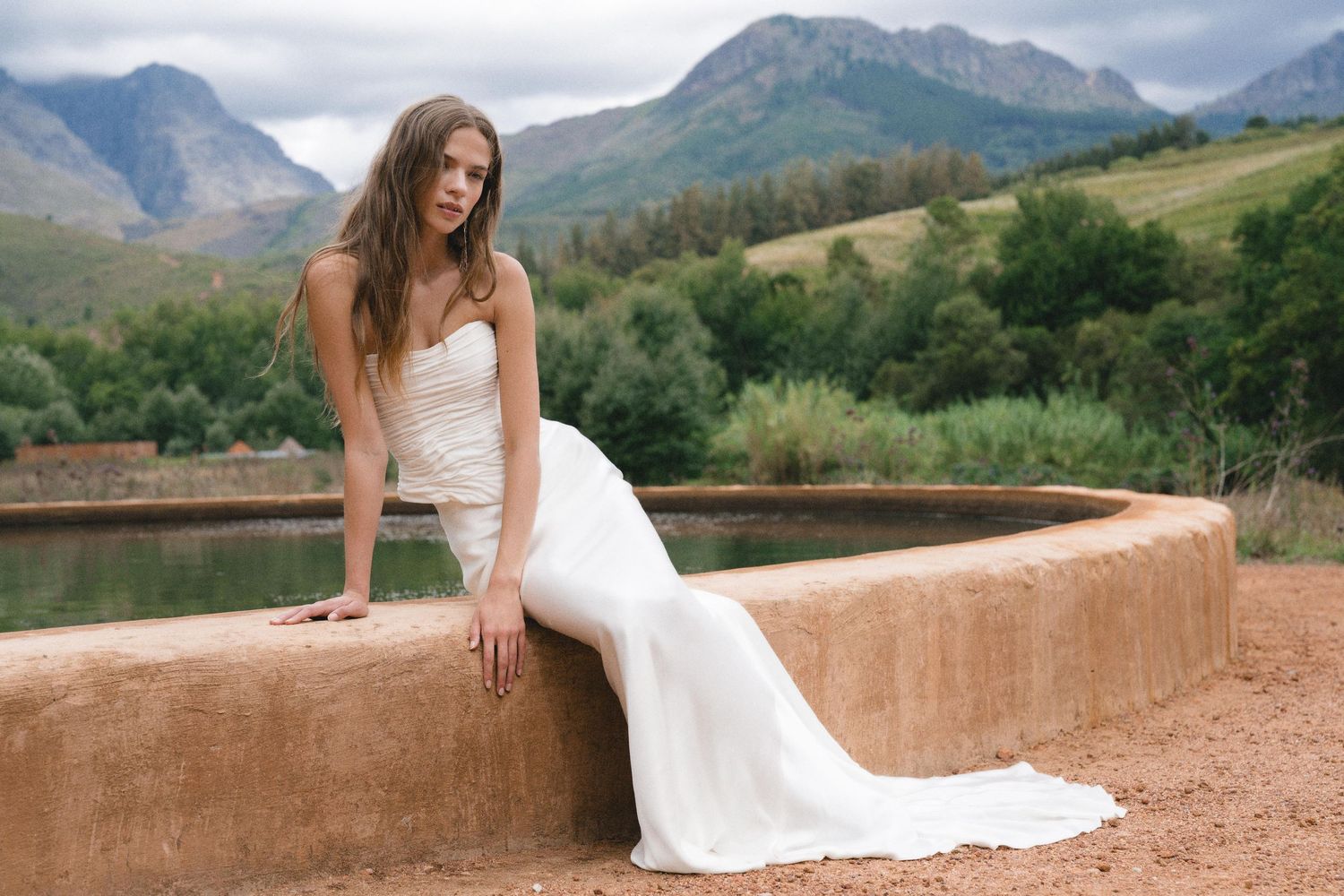 Woman in white strapless wedding gown sits on rustic stone wall with mountain landscape backdrop.
