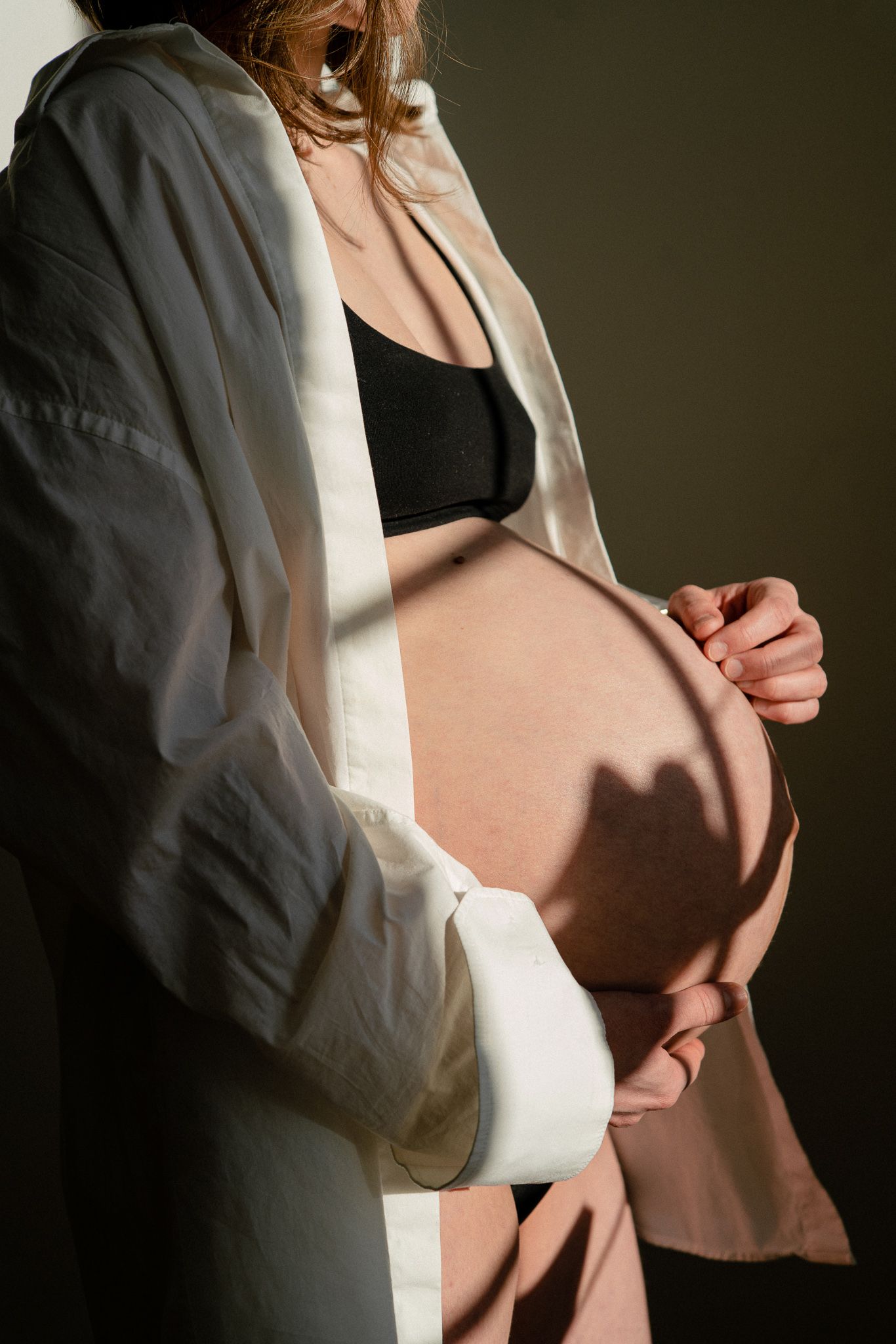Pregnant woman in black bralette and open white blazer cradling her bare baby bump in warm sunlight.