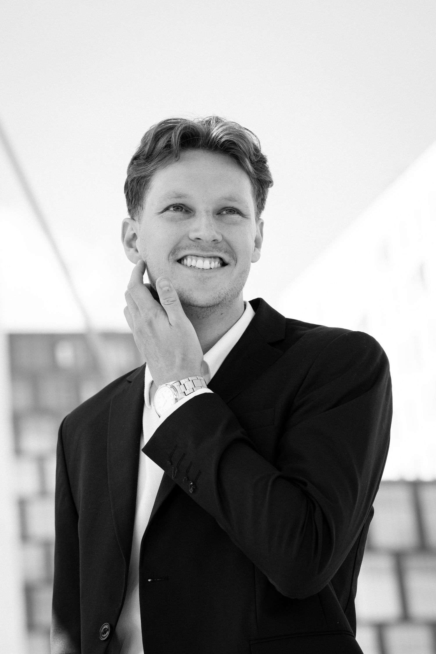 Professional black and white portrait of someone in a dark suit smiling with hand on cheek.