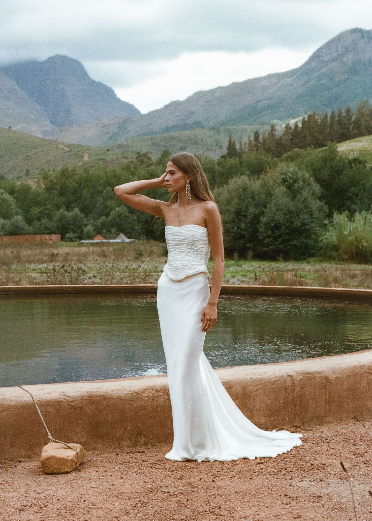 Bride in strapless white gown with draped bodice stands by a pond with mountain backdrop.