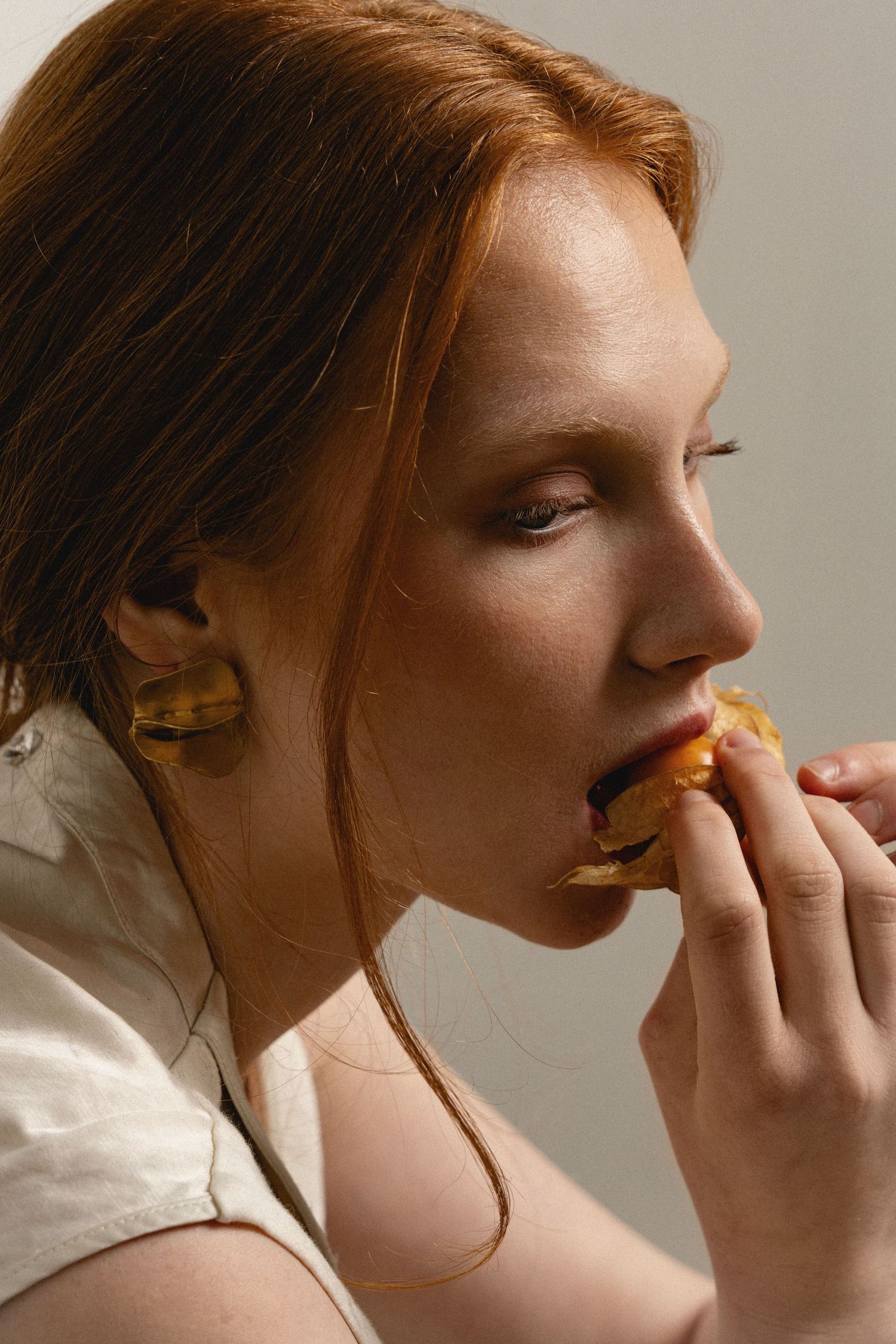 A young redhead woman with freckles eating a pastry, shown in close-up profile view against a neutral background.