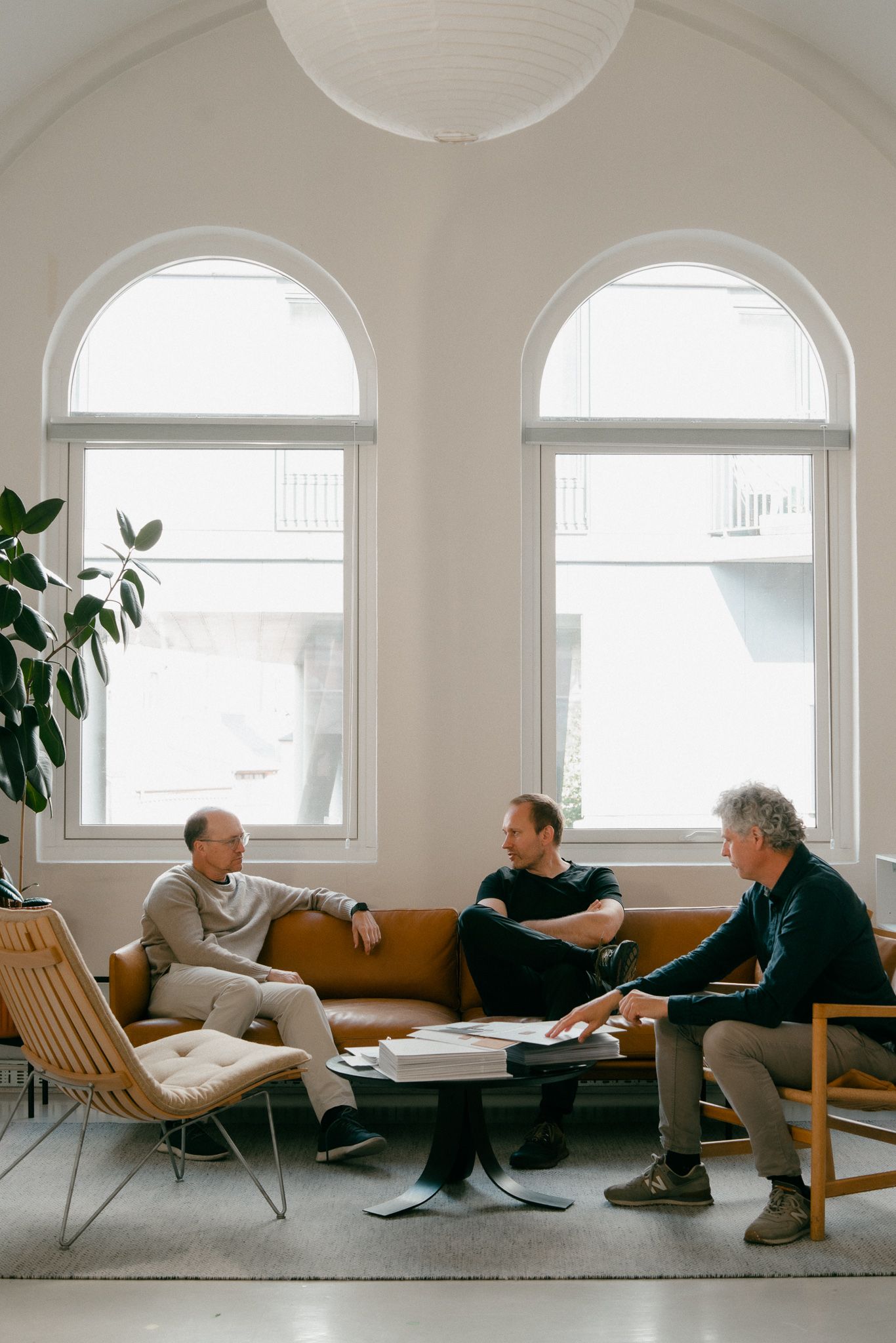 Three people sit in modern lounge chairs by arched windows in a bright minimalist room having a conversation.