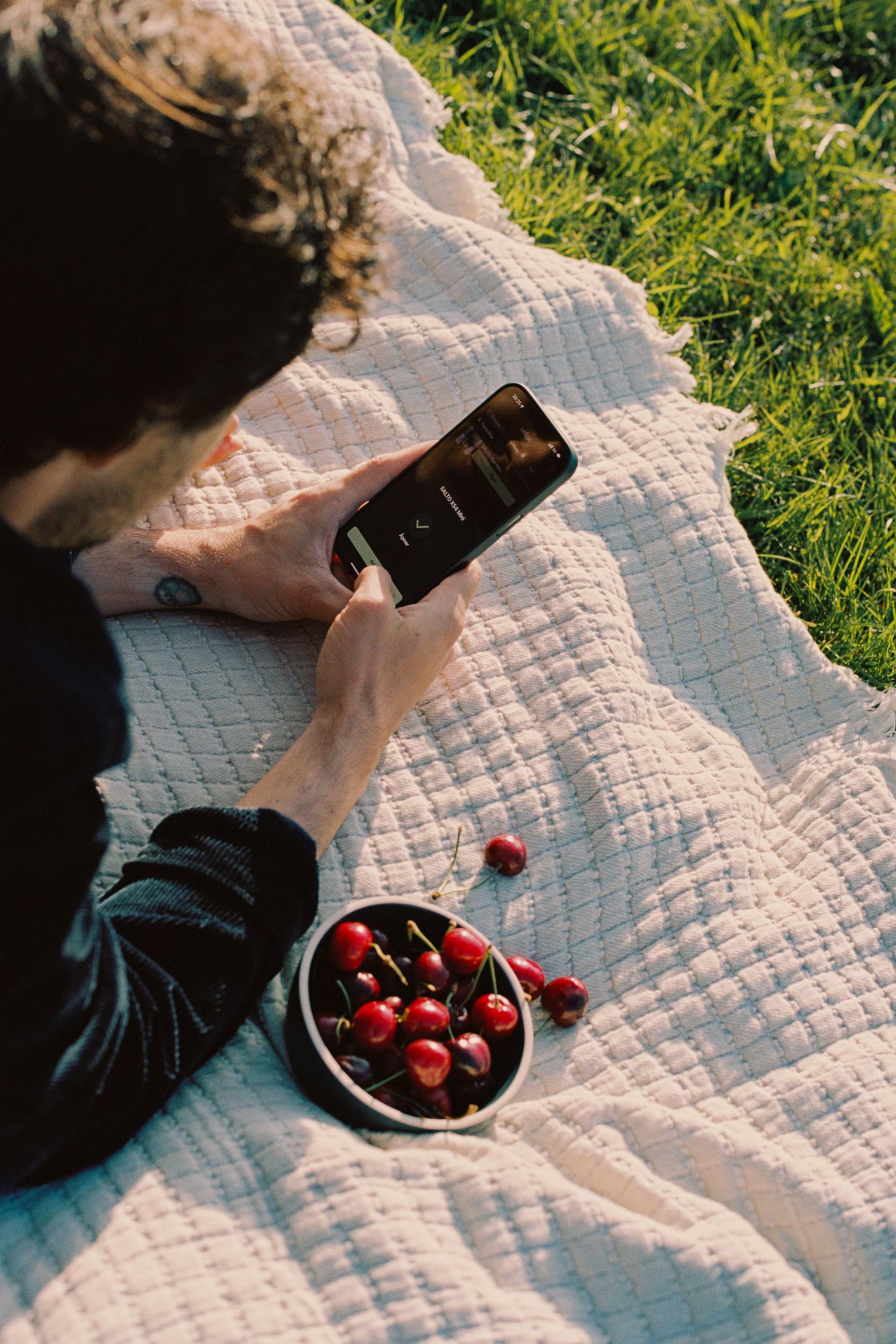 A person lounges on a white quilted blanket in grass while enjoying fresh cherries and a dark beverage.