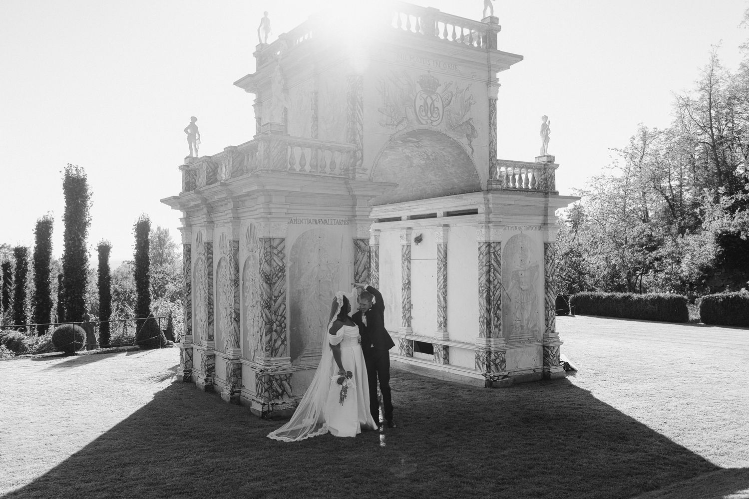 A bride and groom pose for romantic wedding photos in front of a stately white mansion with sun flare in the background.