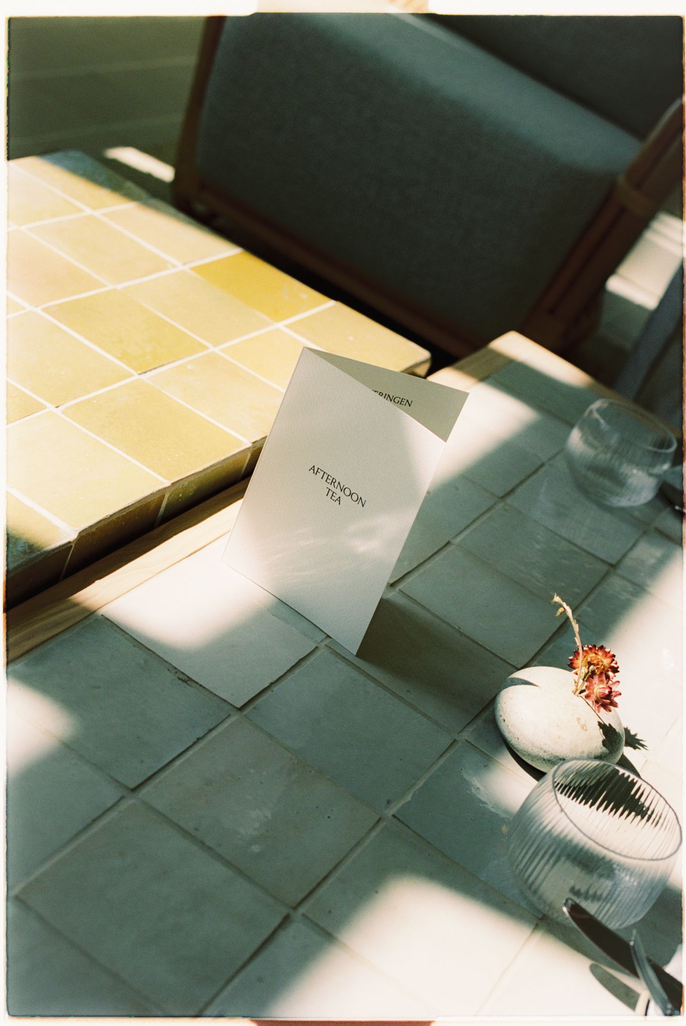 Afternoon tea menu card on tiled table with floral arrangement and glassware in warm sunlight.