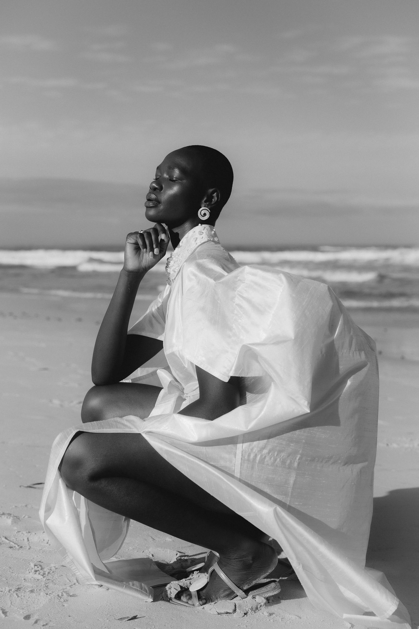 Black and white photo of a woman in flowing white fabric posed gracefully on a sandy beach with ocean waves behind her.