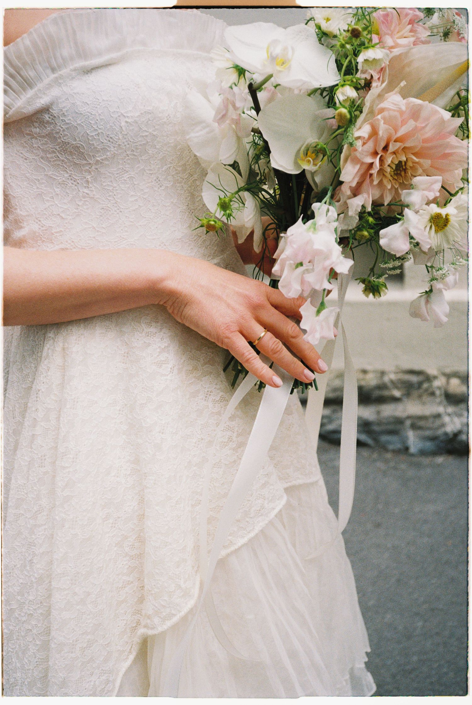 Bride in lace wedding dress holding elegant blush and white floral bouquet with ribbon, showcasing delicate ring on finger.