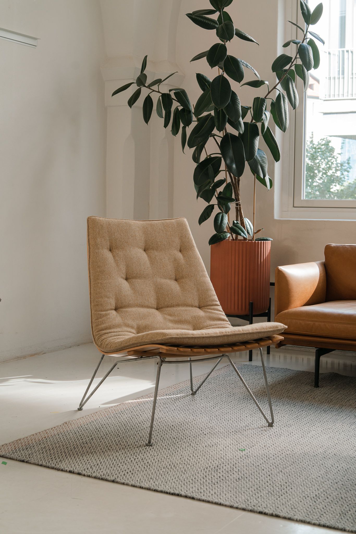 Modern tan leather lounge chair with metal hairpin legs sits on a gray rug next to potted plants in a bright room.