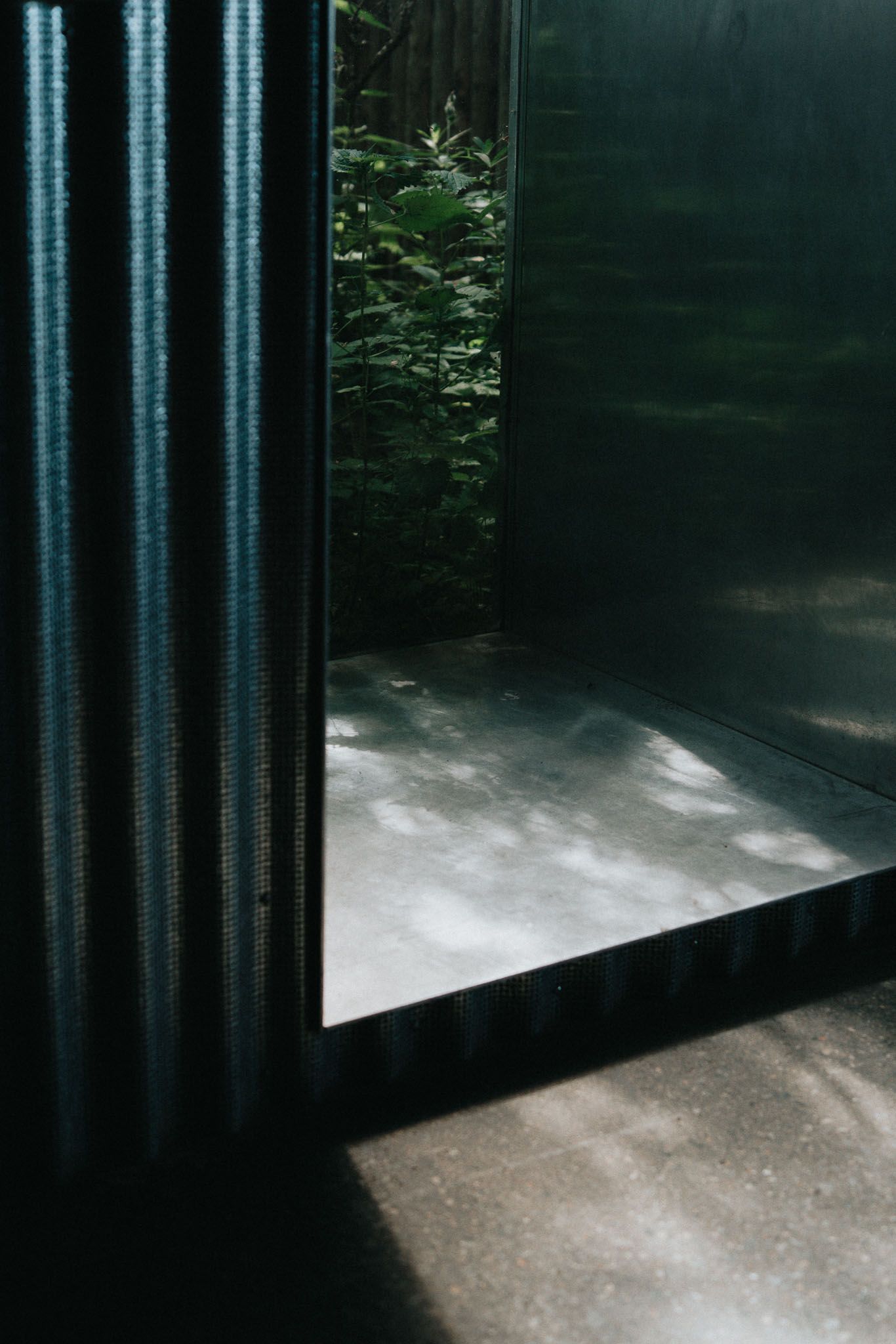 Dramatic interior corner with reflective surfaces and glimpse of greenery through dark architectural elements.