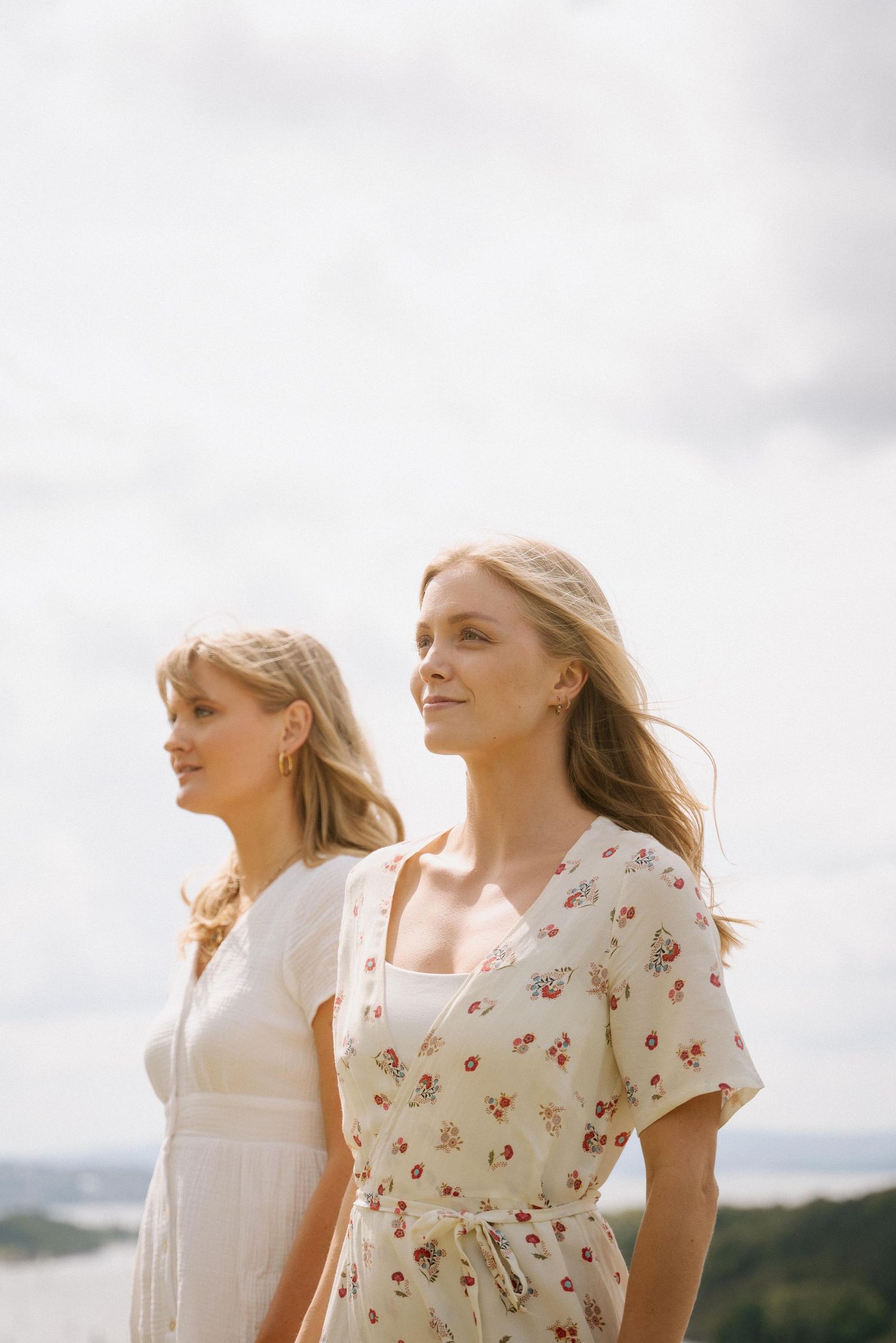 Two women in summer dresses standing outdoors against cloudy sky with scenic view.