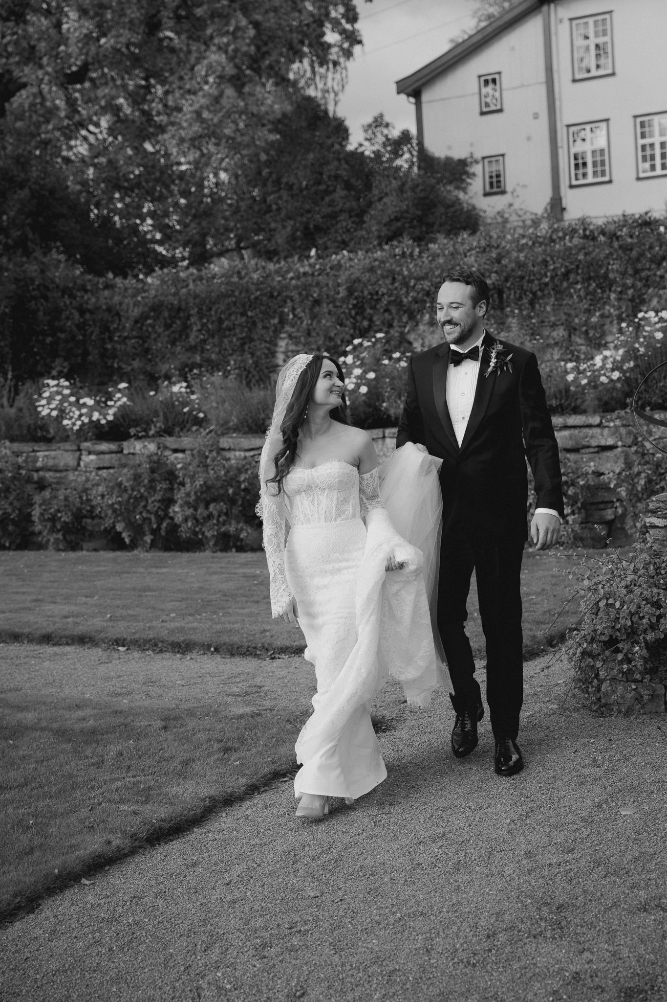 Black and white wedding photos show couple walking together through a garden path in formal attire.