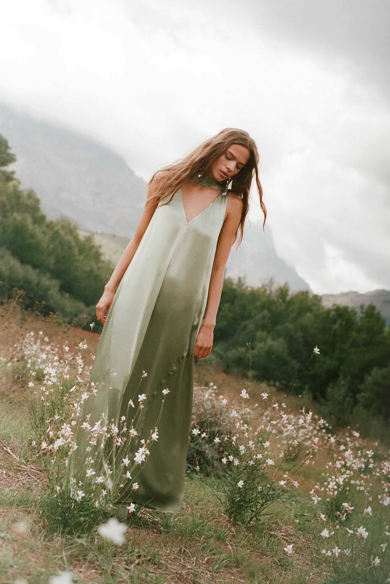 Woman in sage green maxi dress standing among white wildflowers with misty mountains in background.