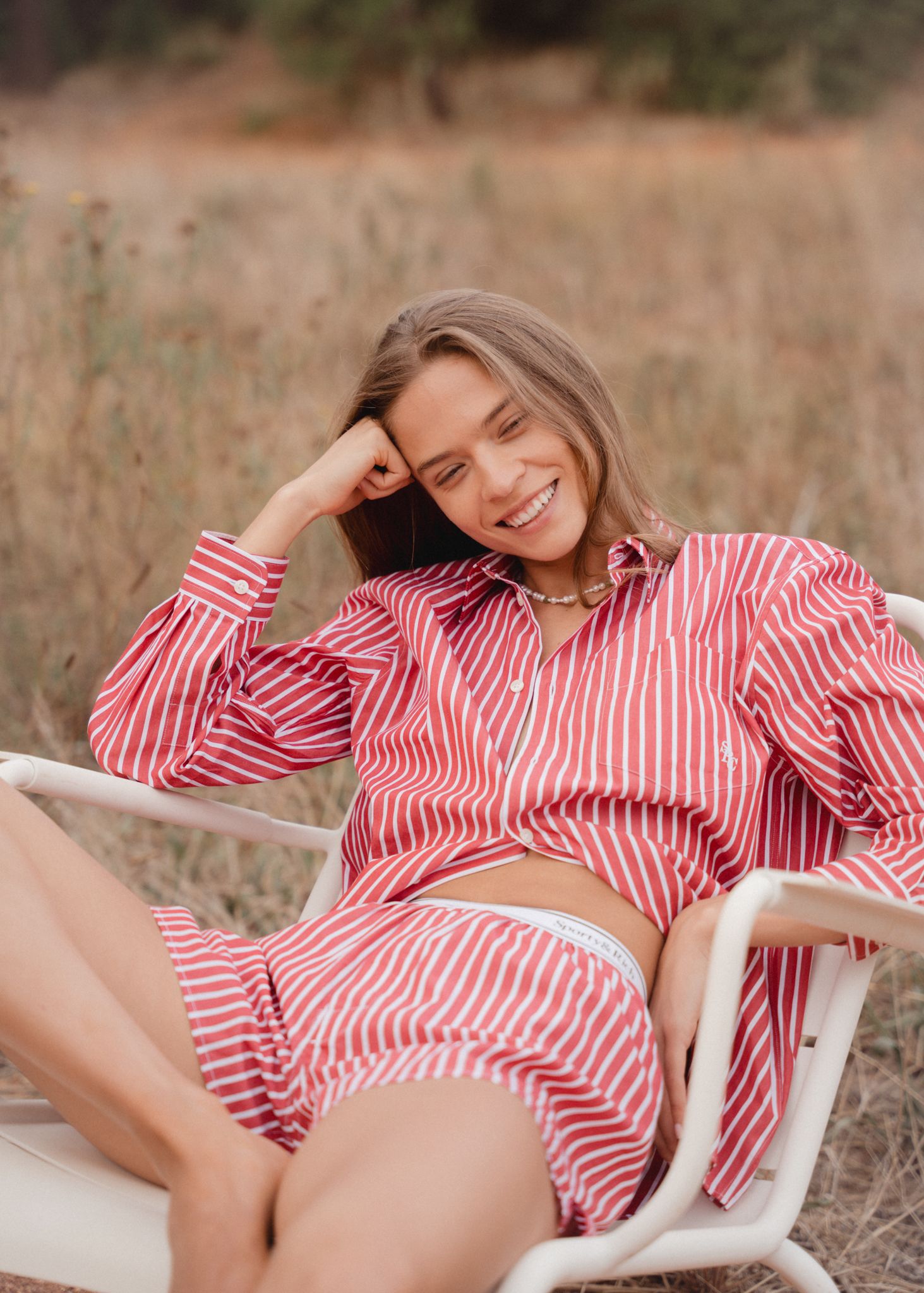 Woman smiling in red striped shirt and shorts, relaxing in white chair outdoors in a natural field setting.