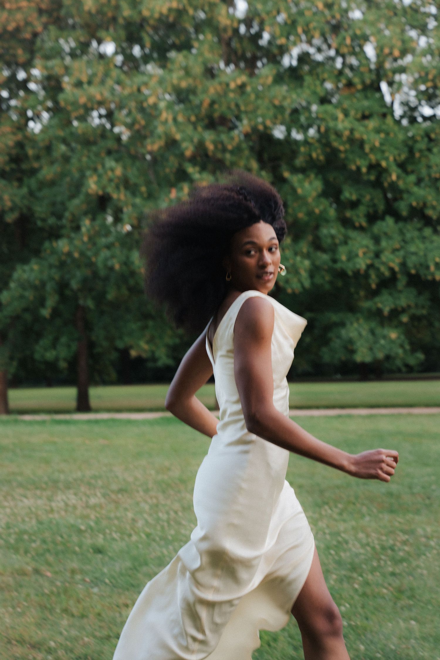A young Black woman with an afro runs through a green park in a flowing white sleeveless dress, glancing back.