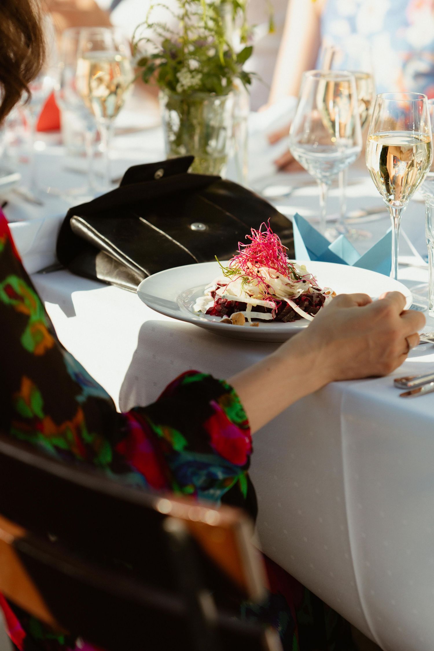 Woman in floral dress dining at elegant table with gourmet salad plate and champagne glasses in sunlight.