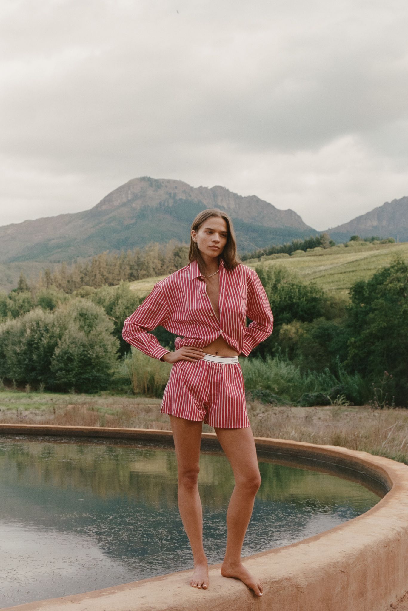 Woman in pink striped matching set stands barefoot by a pool with mountain views in the background.
