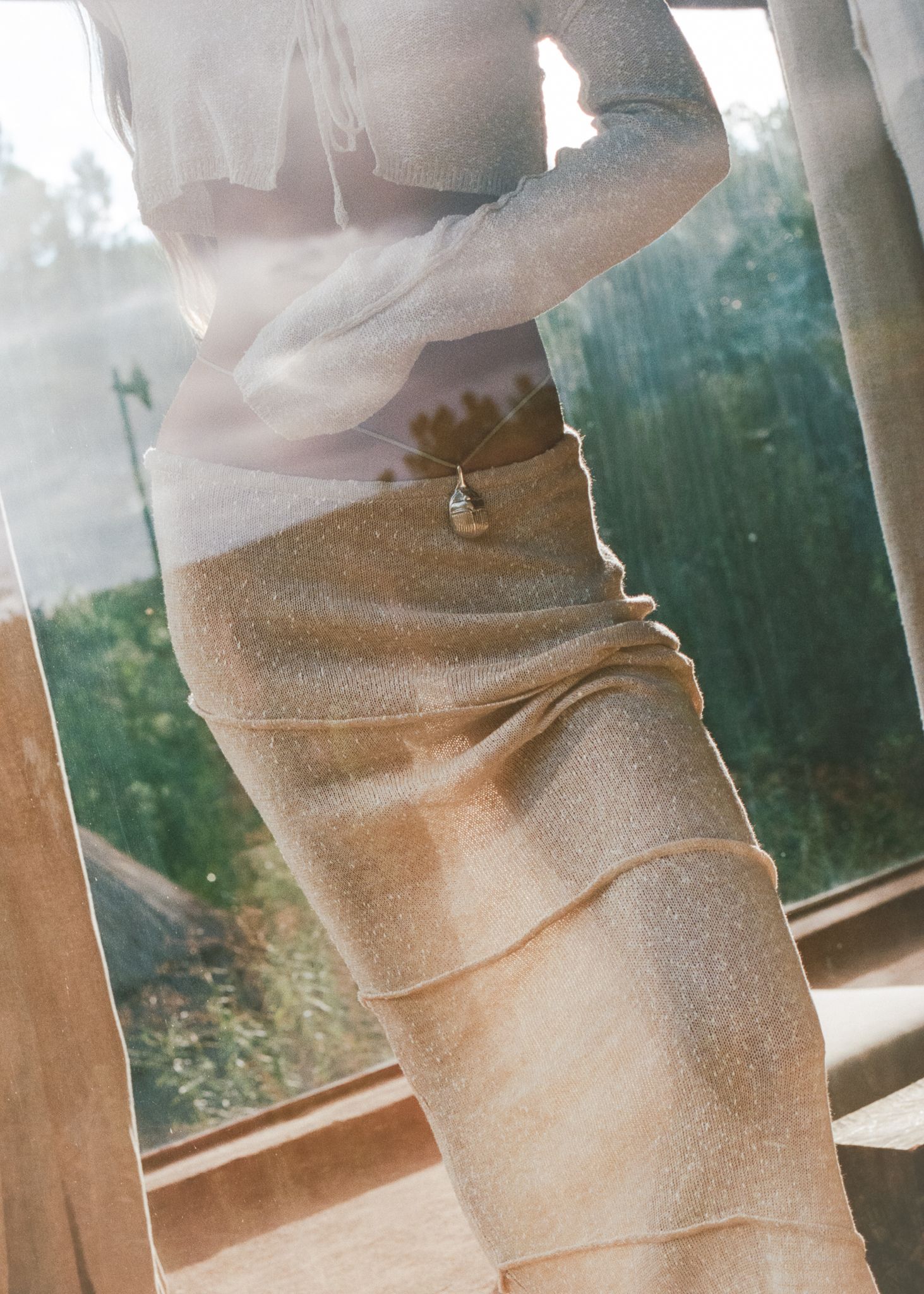 Woman wearing a sheer white top and gold metallic midi skirt with pendant necklace, standing by a sunlit window.