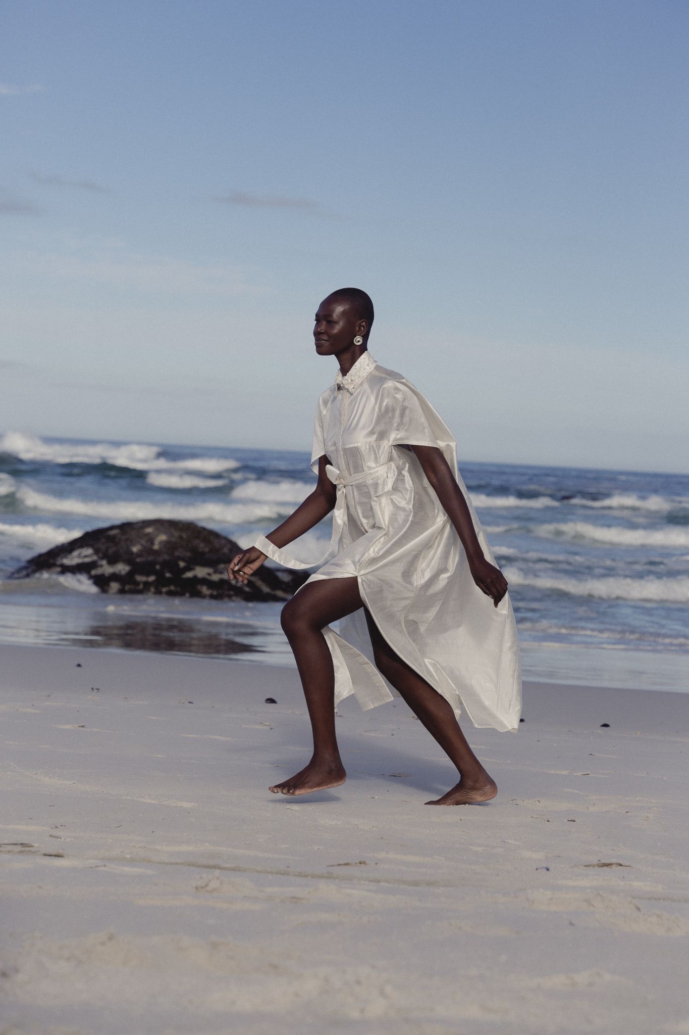 A tall Black woman in a flowing white dress walks barefoot on a sandy beach with ocean waves behind her.