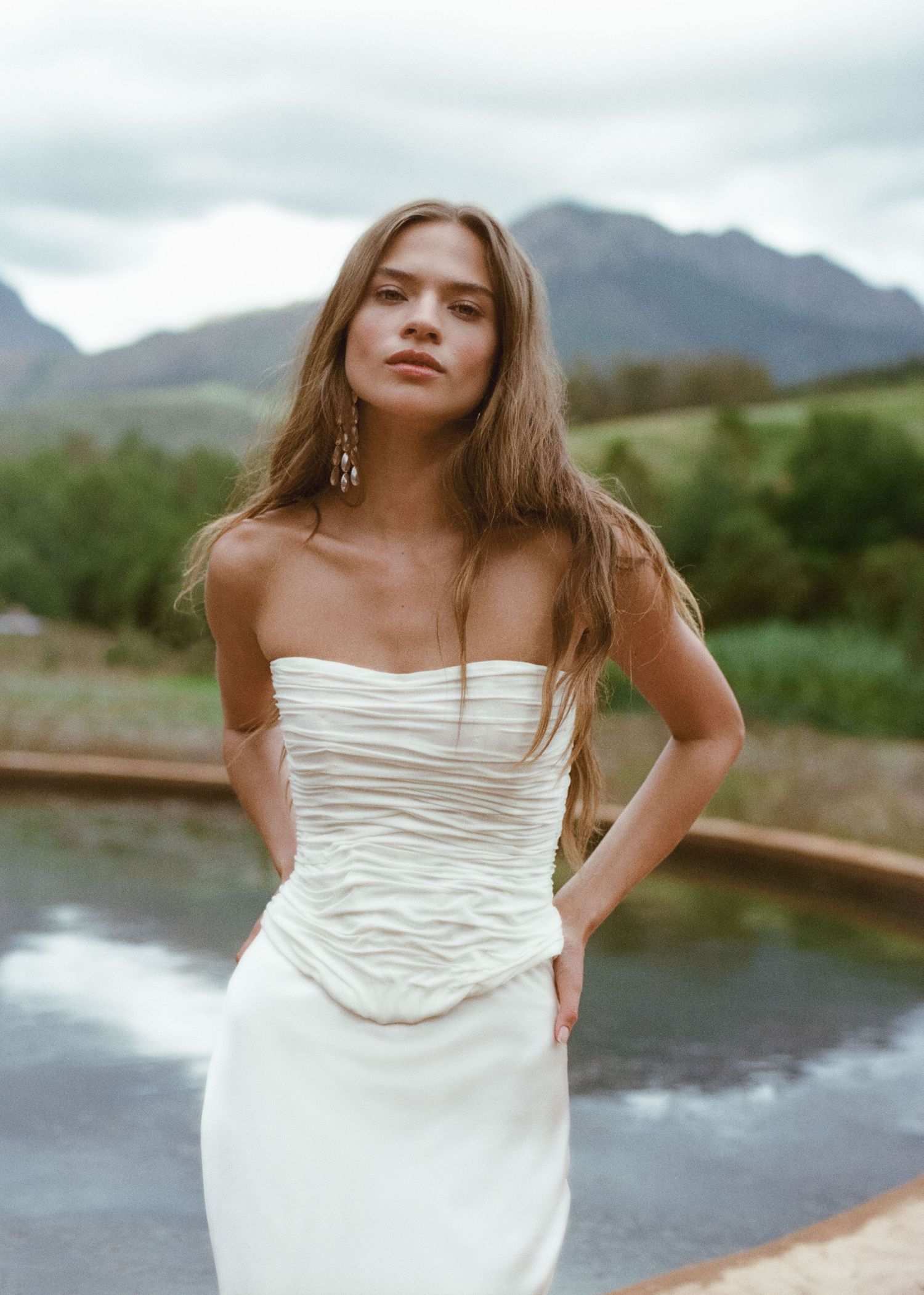 Woman in white strapless ruched gown poses outdoors with mountain backdrop and overcast sky.