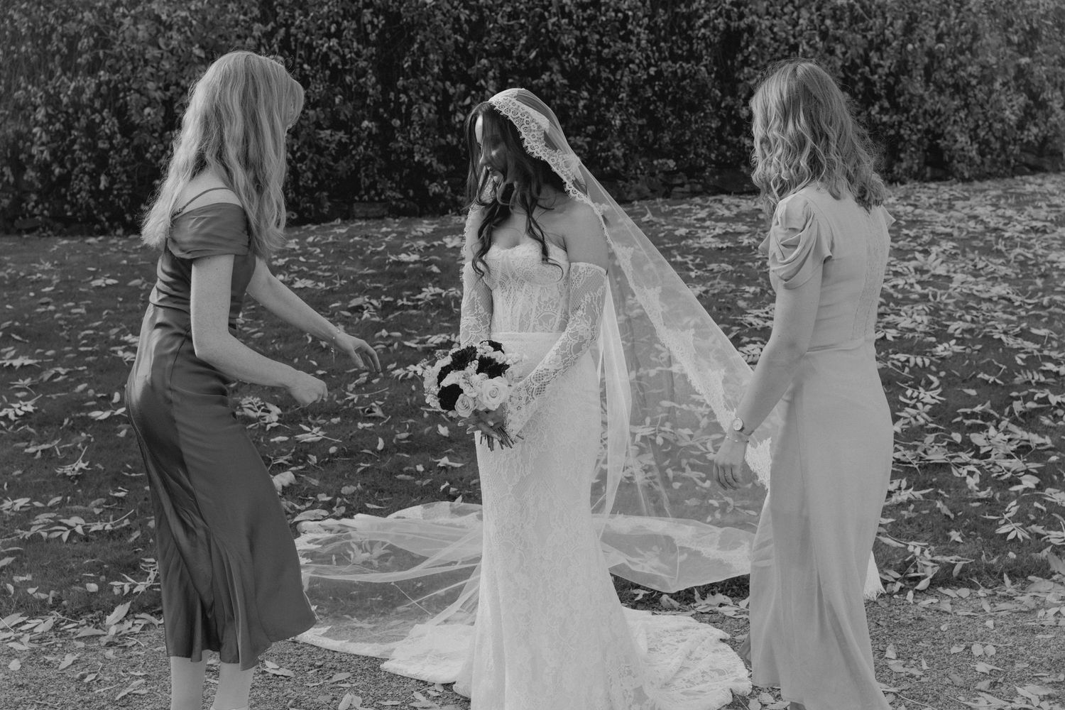 Black and white photo of a bride in lace gown with two bridesmaids adjusting her veil outdoors.