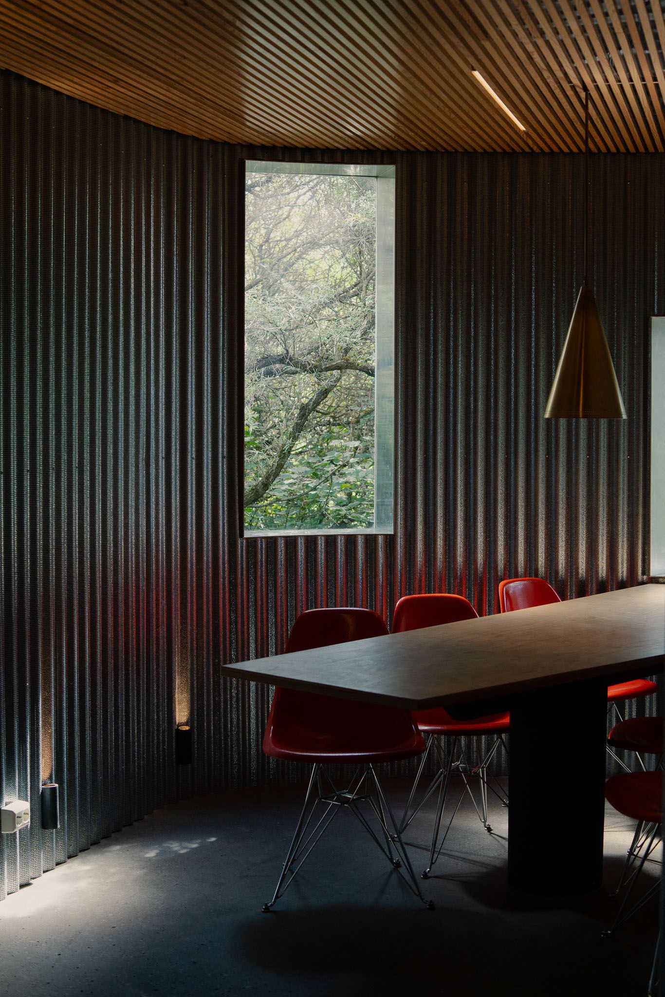 Modern dining room with dark wood paneling, red chairs, and tall window framing outdoor trees.