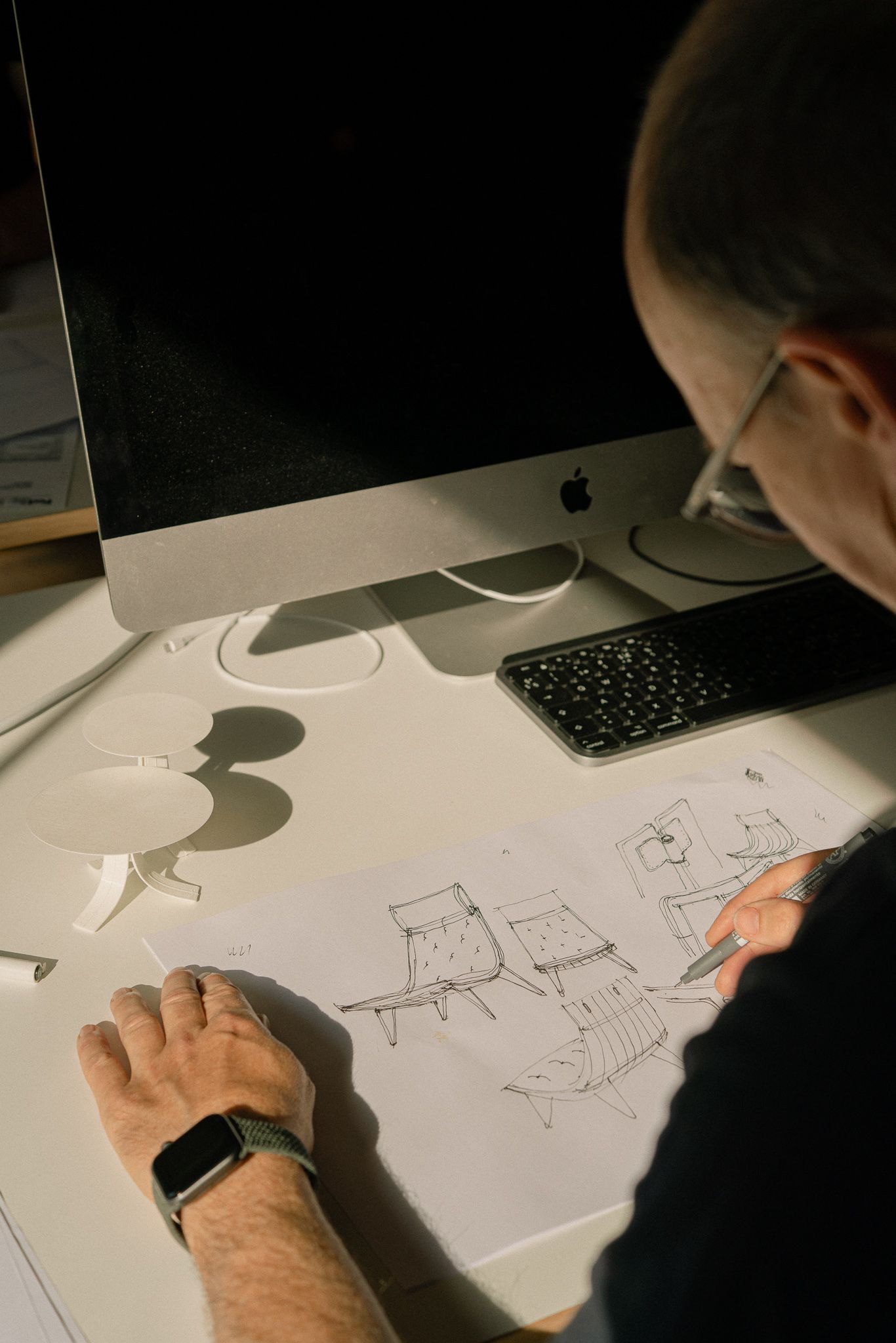 An artist sketches furniture designs on white paper at a desk with a computer monitor casting shadows.