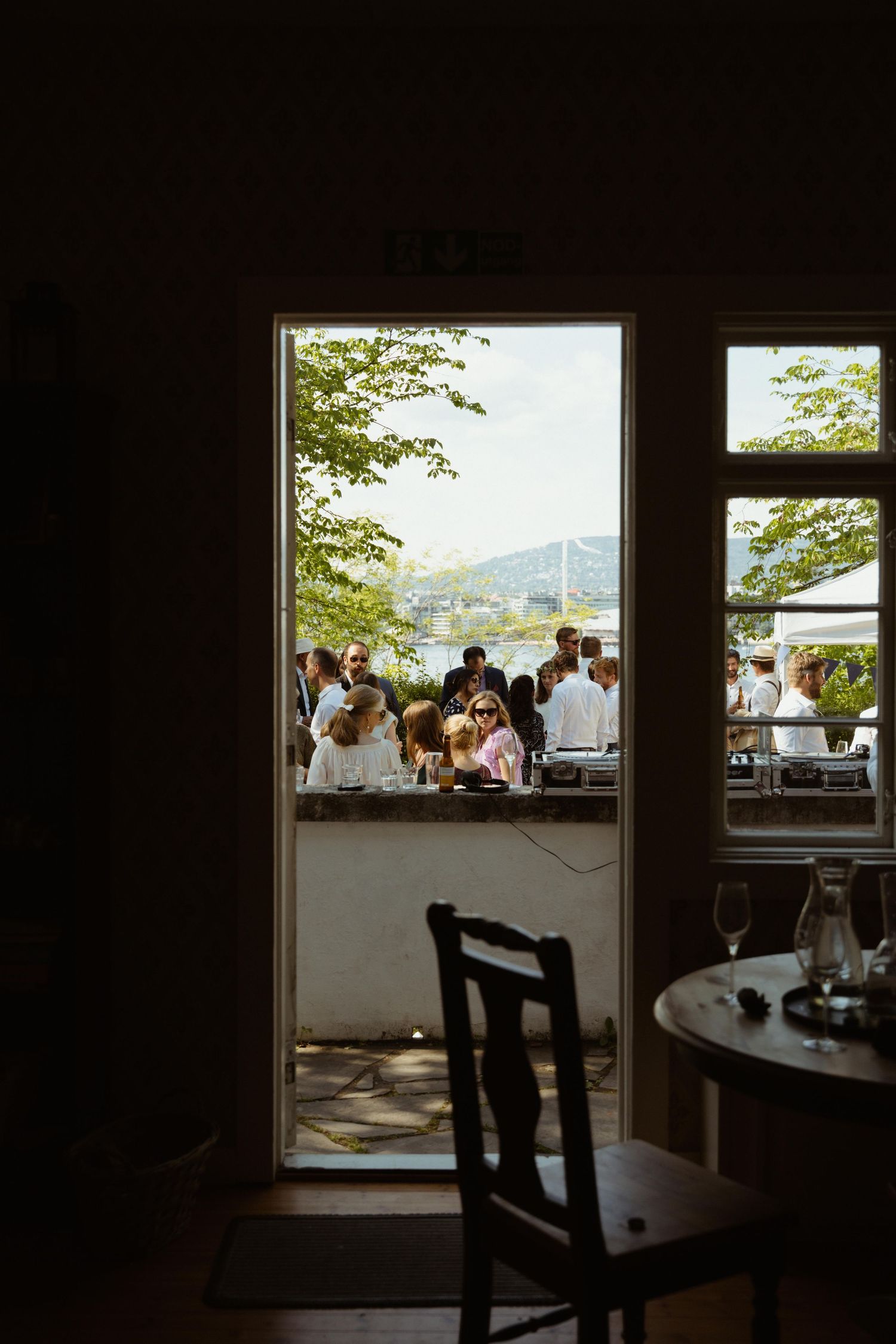 View through open door to outdoor gathering with people dining al fresco near waterfront with trees and bridge.