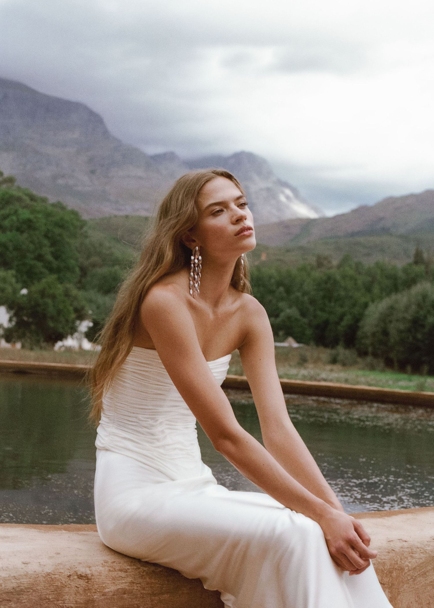 Woman in white strapless dress sits by mountain lake, wearing long earrings, with lush green trees and cloudy peaks behind.