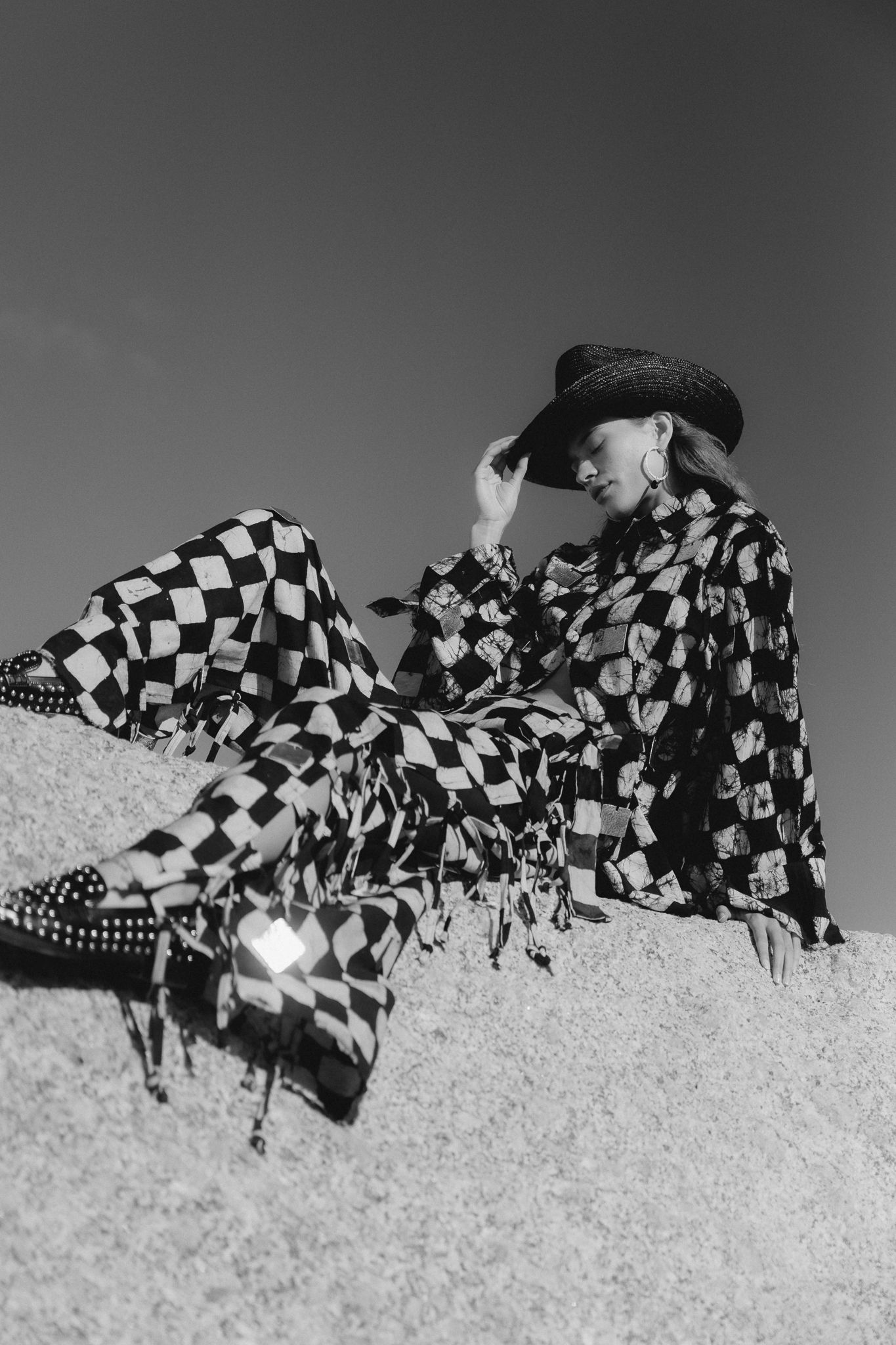Black and white photo of a woman in checkered outfit and cowboy hat sitting on sandy dunes.