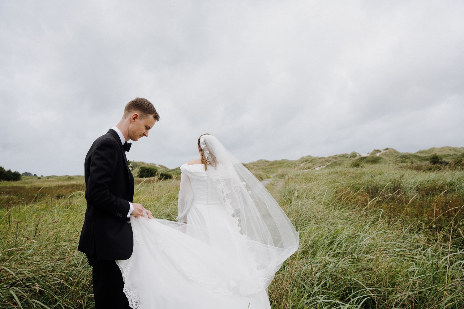 A bride and groom share a romantic moment in a windswept field of tall grass under cloudy skies.