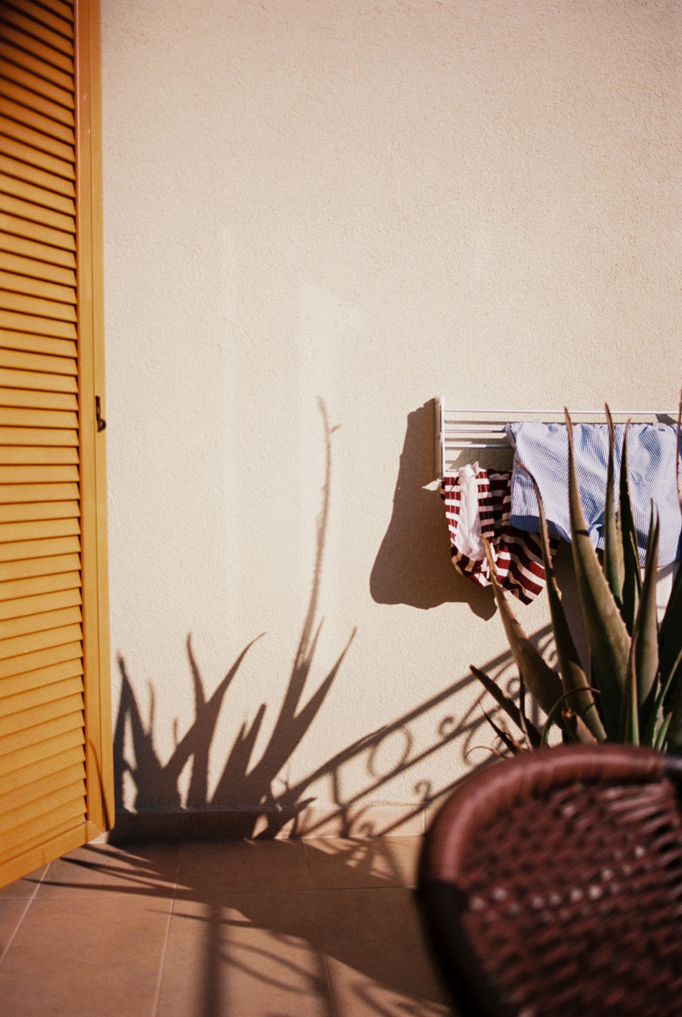 Laundry drying on a line casts shadows against a beige wall next to wooden shutters and a potted plant.