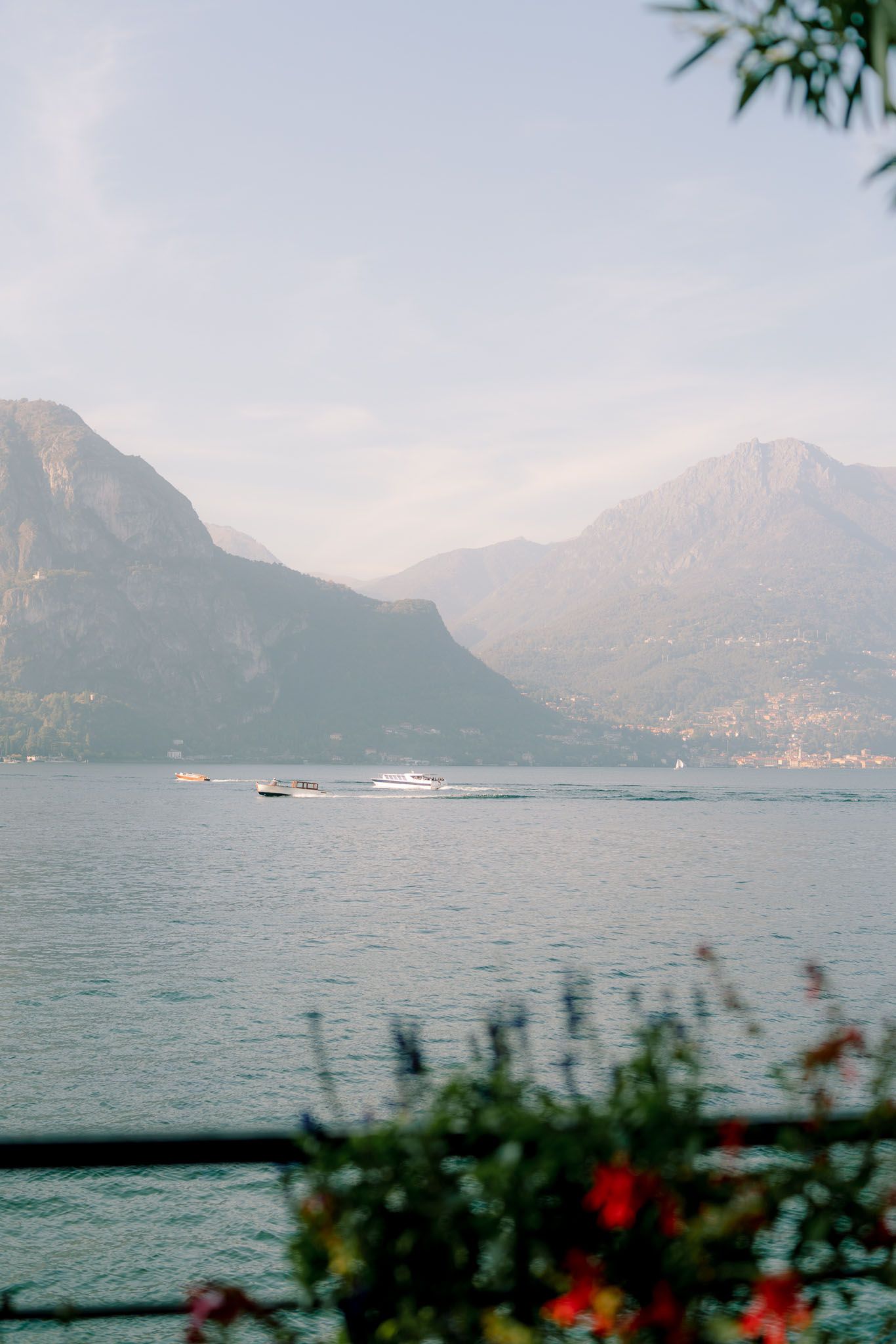 Misty mountain landscape overlooking Lake Como with boats on the water and colorful flowers in the foreground.