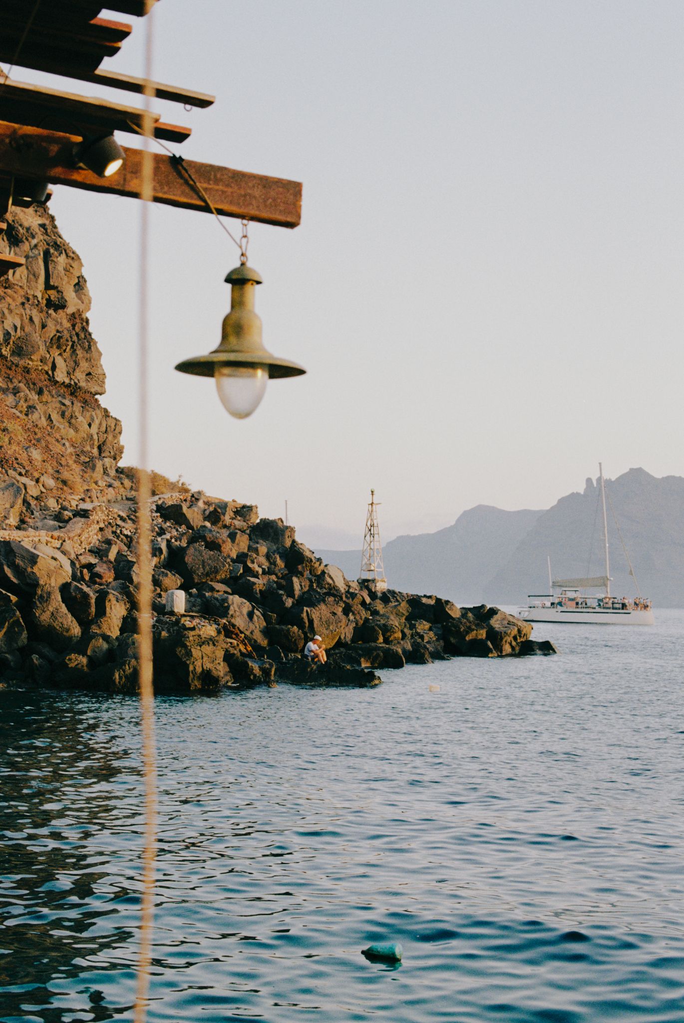 A rustic lamp hangs over Mediterranean waters with rocky coastline and sailboat silhouetted against mountain backdrop.