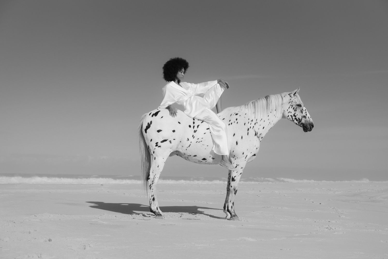 Black and white photo of a woman in a flowing white dress riding a spotted Appaloosa horse on a sandy desert landscape.