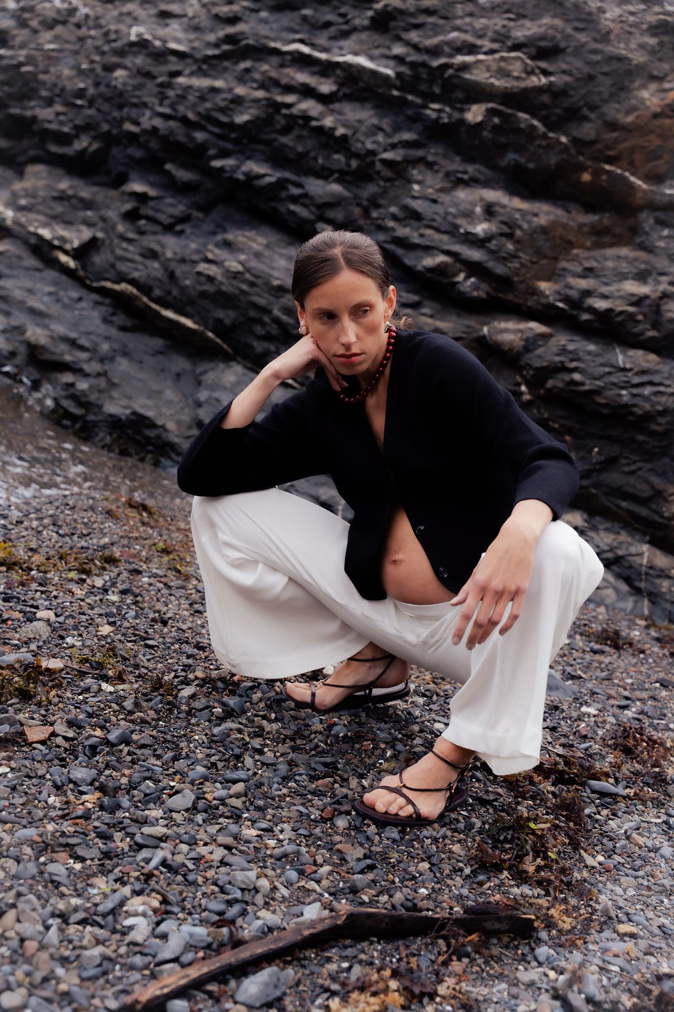 Woman in black top and white pants crouching on rocky beach with dark stone cliff backdrop.