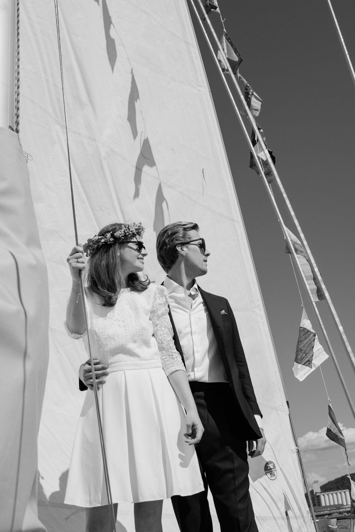 Black and white photo of a bride and groom in sunglasses standing by sailboat masts on their wedding day.