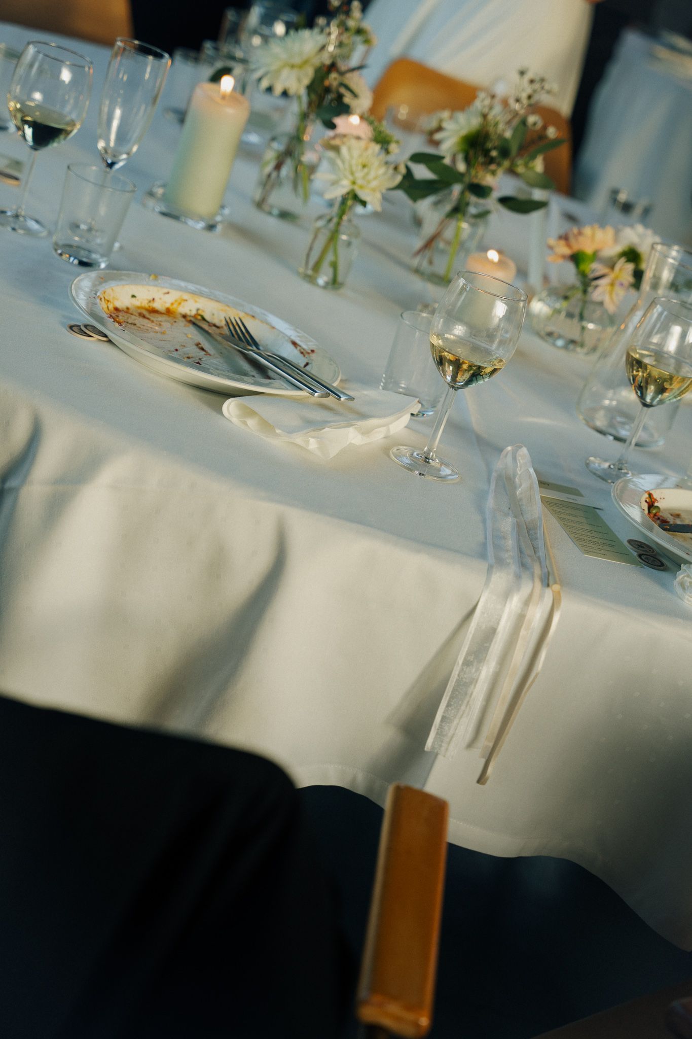 Elegant wedding reception table settings with white linens, wine glasses, and delicate floral centerpieces in soft lighting.