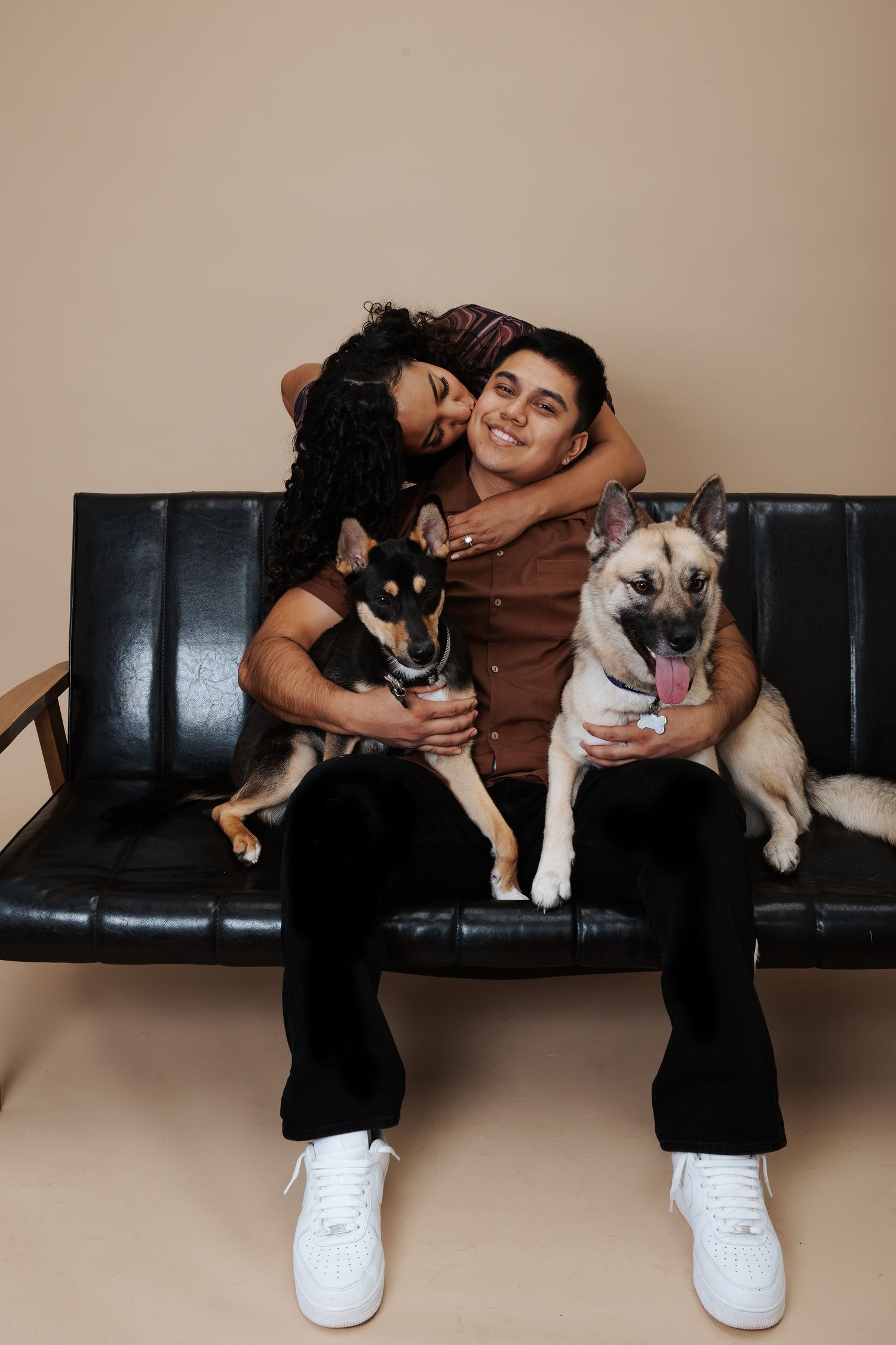 A couple sits on a black leather bench with their two dogs, enjoying a warm embrace against a beige wall.