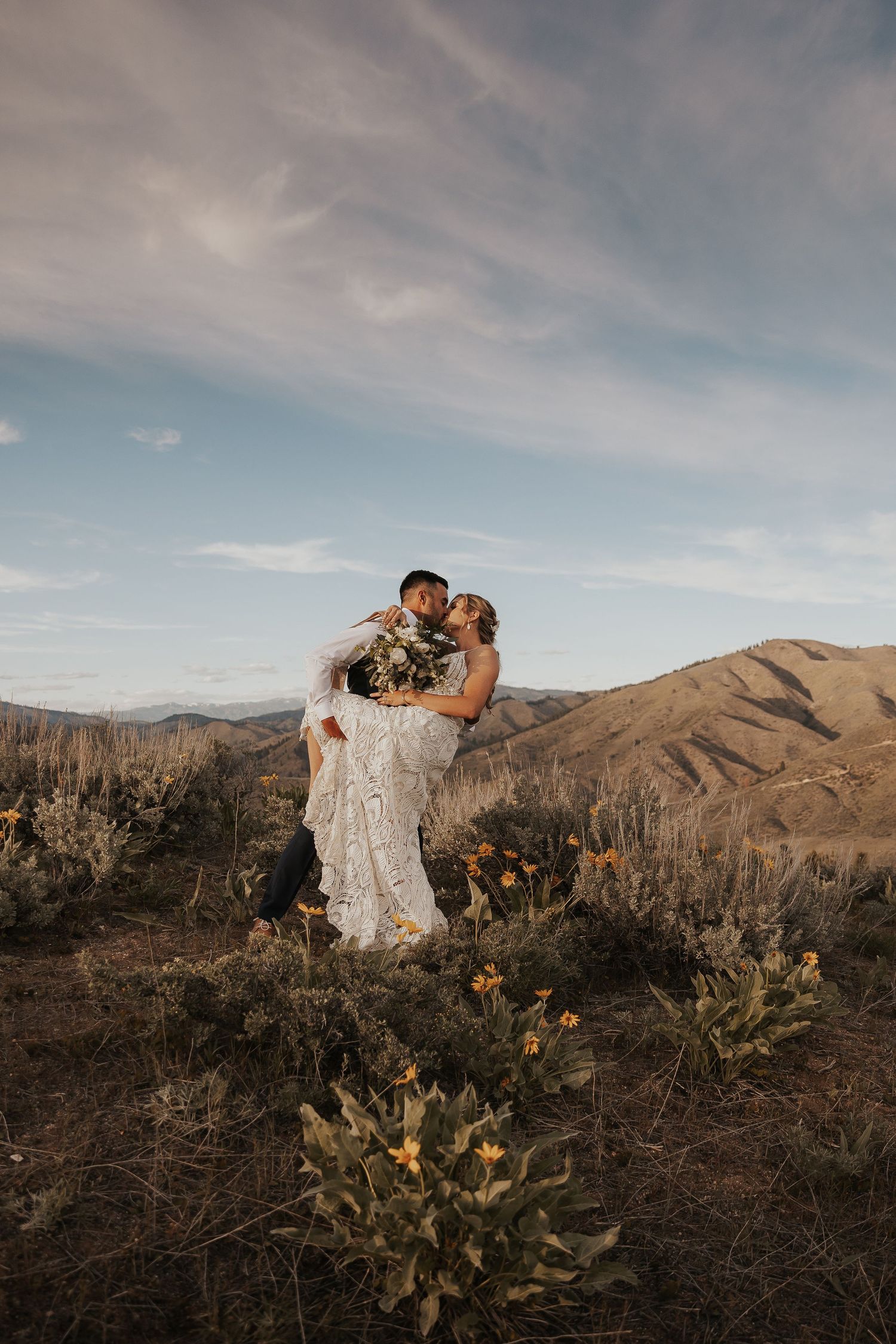 Wedding couple embracing on mountainside at sunset with desert landscape.