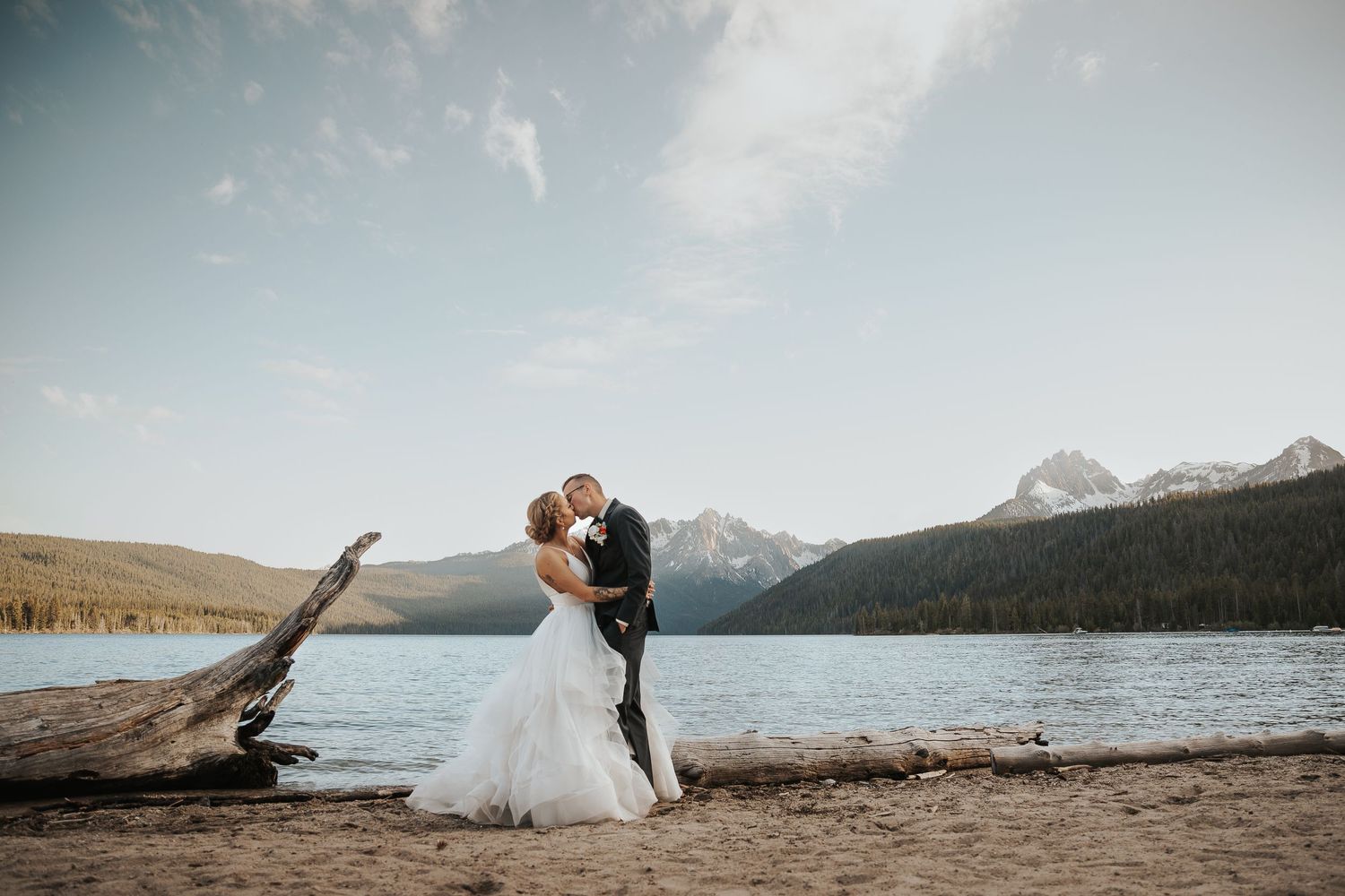 A couple shares a romantic moment by a serene mountain lake with snow-capped peaks in the background at sunset.