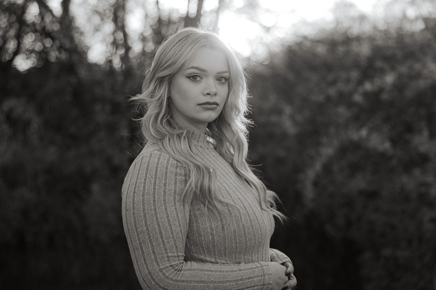 Black and white outdoor portrait of a person with long wavy blonde hair wearing a ribbed sweater with trees in background.